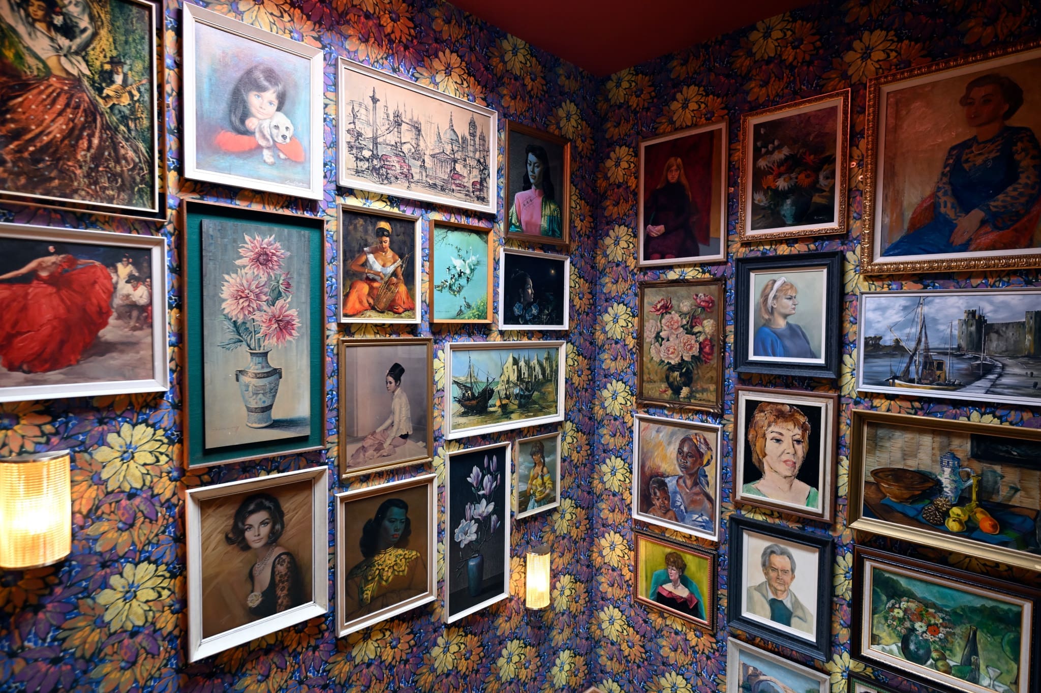 A room with floral wallpaper displays a dense arrangement of framed paintings, from portraits to ships, creating an eclectic fossato-inspired gallery wall. Two wall sconces provide warm lighting to this colorful display.