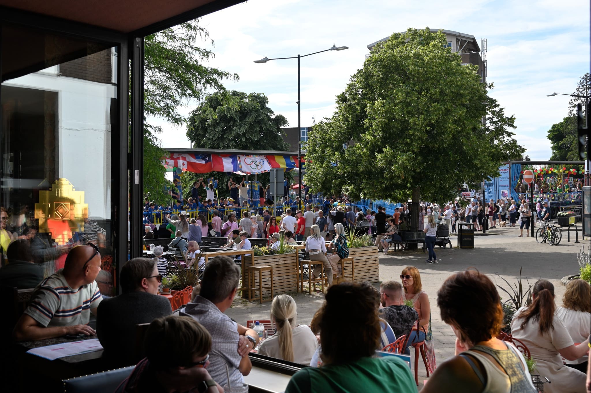 People sit at outdoor tables near a cafe by the fossato, watching a lively street parade with dancers in blue costumes and headdresses. Trees and a festive stage with colorful decorations are visible under a sunny sky.