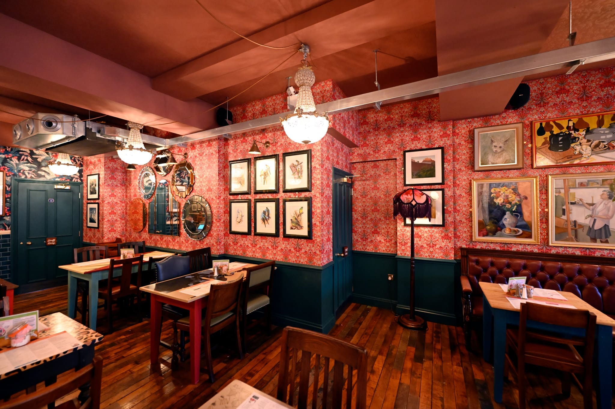 A cozy restaurant interior in Francisco with floral wallpaper, framed art on the walls, vintage chandeliers, wooden tables and chairs, green paneling, and a wooden floor. The eclectic space feels uniquely inviting.