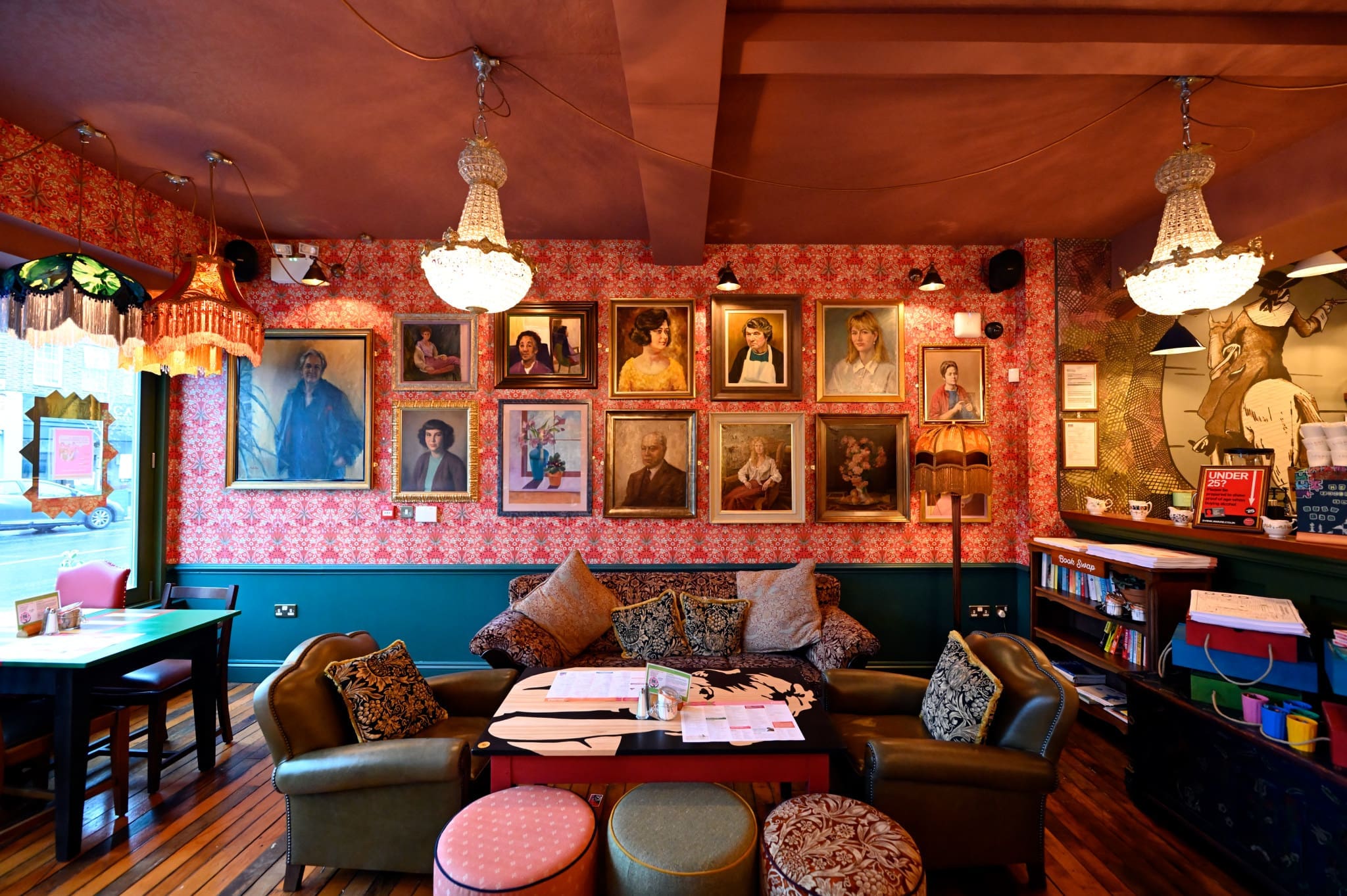 A cozy cafe with floral wallpaper, vintage portraits—including one of Francisco—chandeliers, a sofa, armchairs, and round ottomans around a low table; bookshelves and a window add to the eclectic, inviting atmosphere.