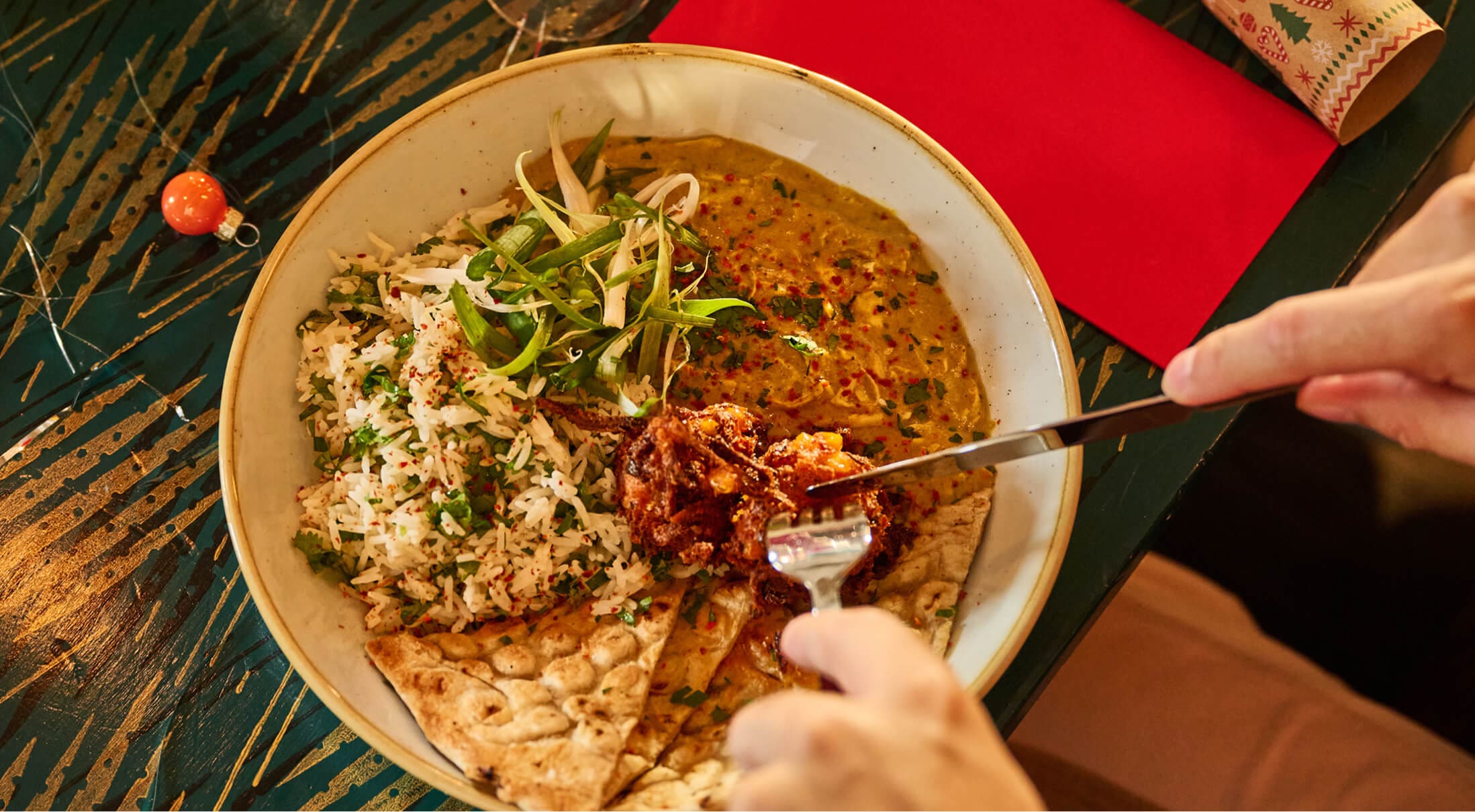 A bowl of rice, flatbread, curry, and lentil stew garnished with fresh greens on a patterned table. A hand is using a fork and knife to cut a piece of food from the dish.