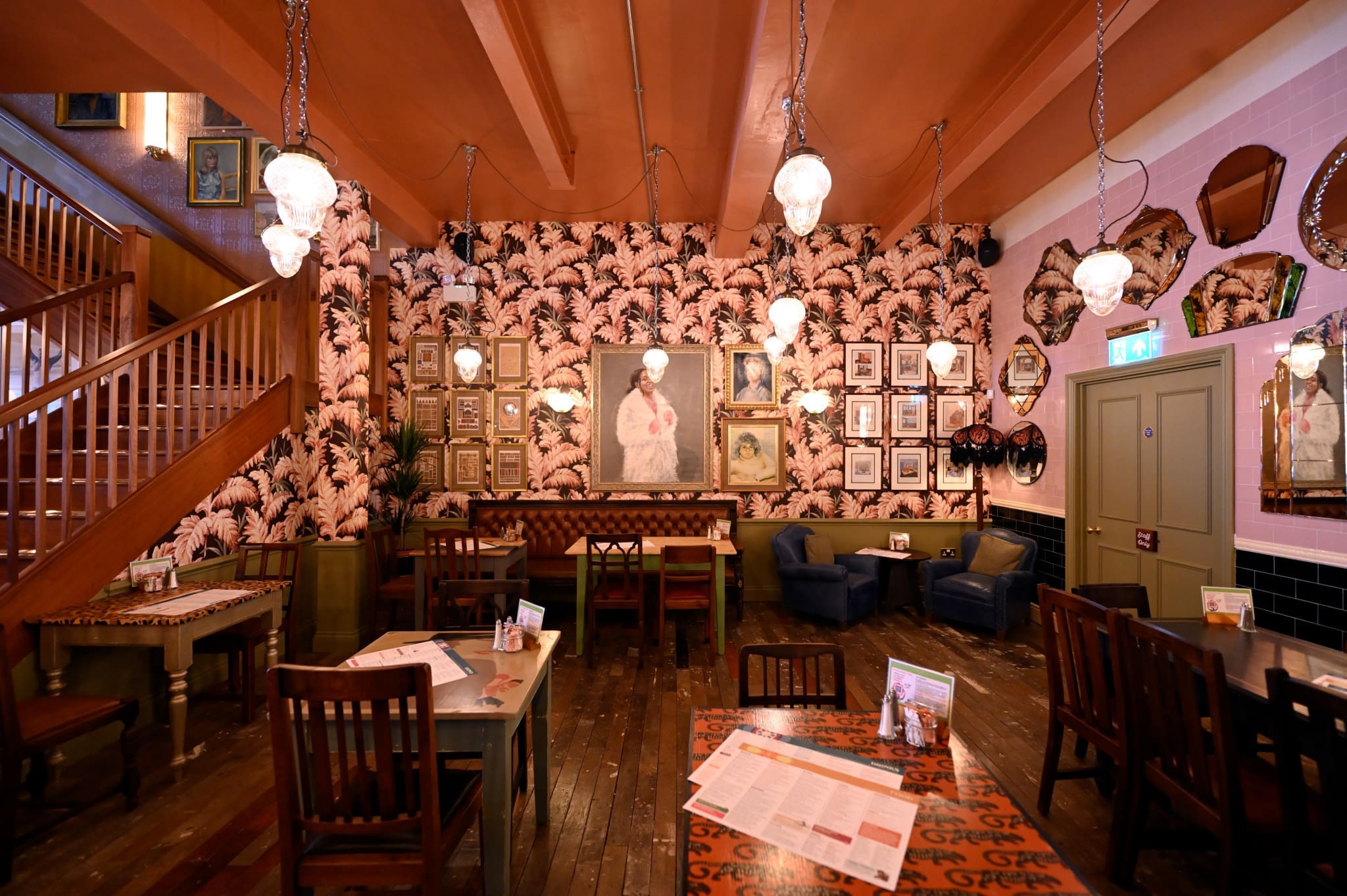 A cozy, vintage-style cafe with patterned wallpaper, framed art, hanging pendant lights, wooden tables and chairs, and a staircase leading to an upper Granero-inspired level. The ambiance is warm and inviting.