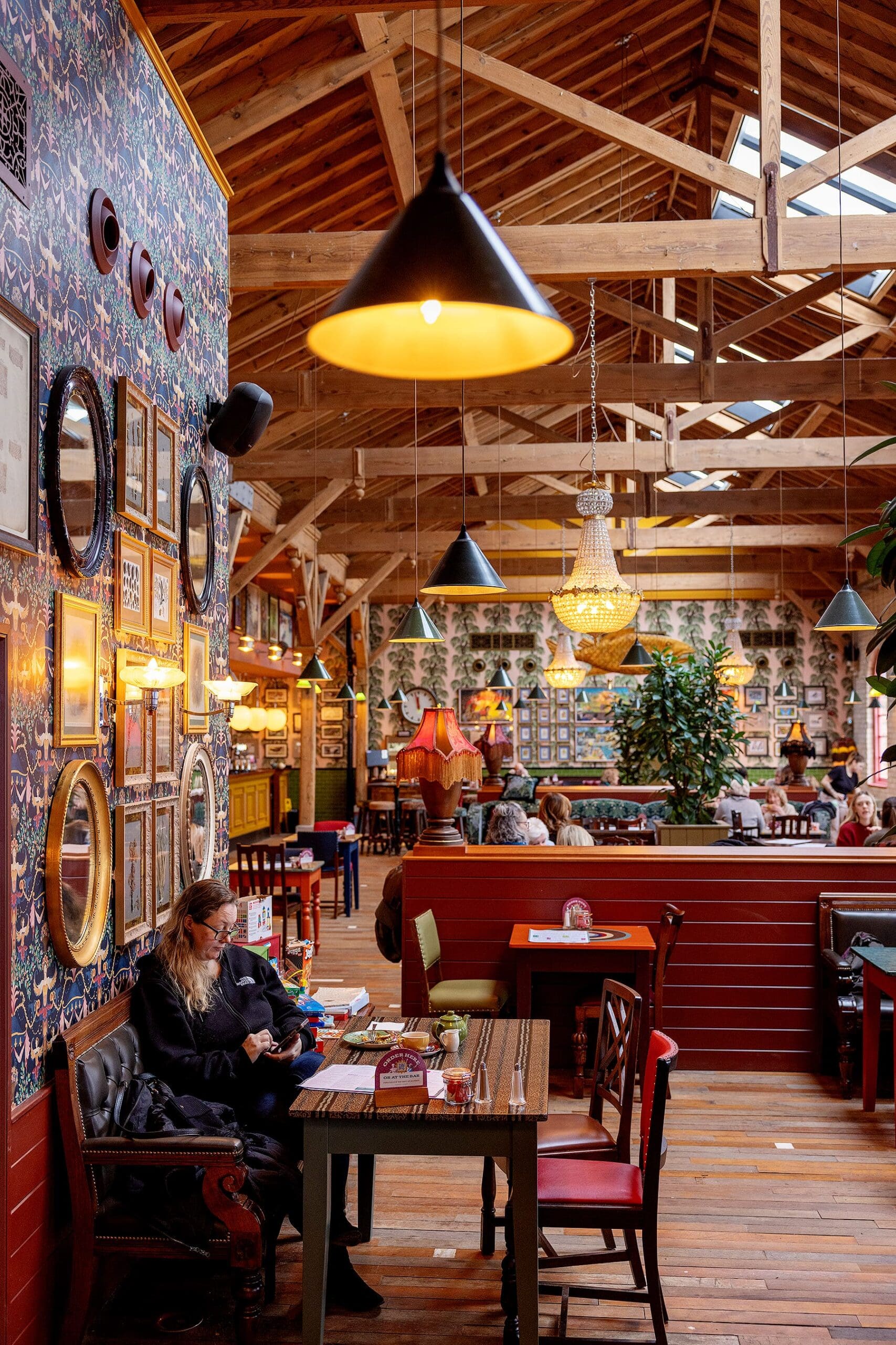 A cozy, eclectic café with wooden beams, warm hanging lights, and a wall decorated with framed pictures. People sit at tables and booths, some chatting, while a woman works on a laptop near the foreground.