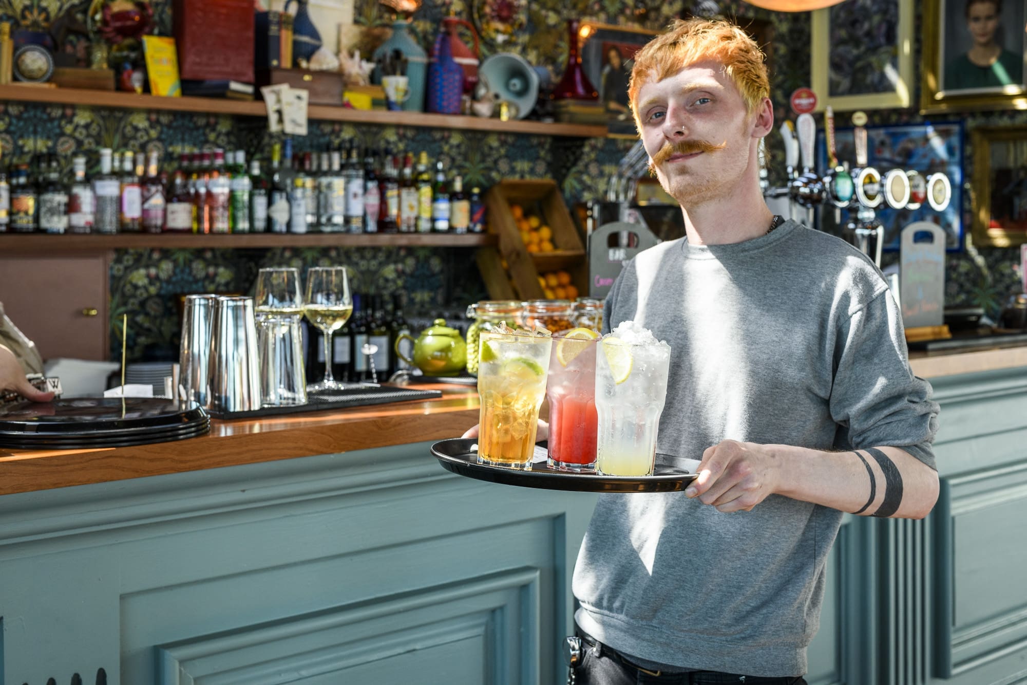 A server with red hair and a mustache holds a tray with colorful cocktails and lemonade in a bright, stylish bar with a teal counter—showcasing vibrant hospitality careers amid shelves of bottles in the background.