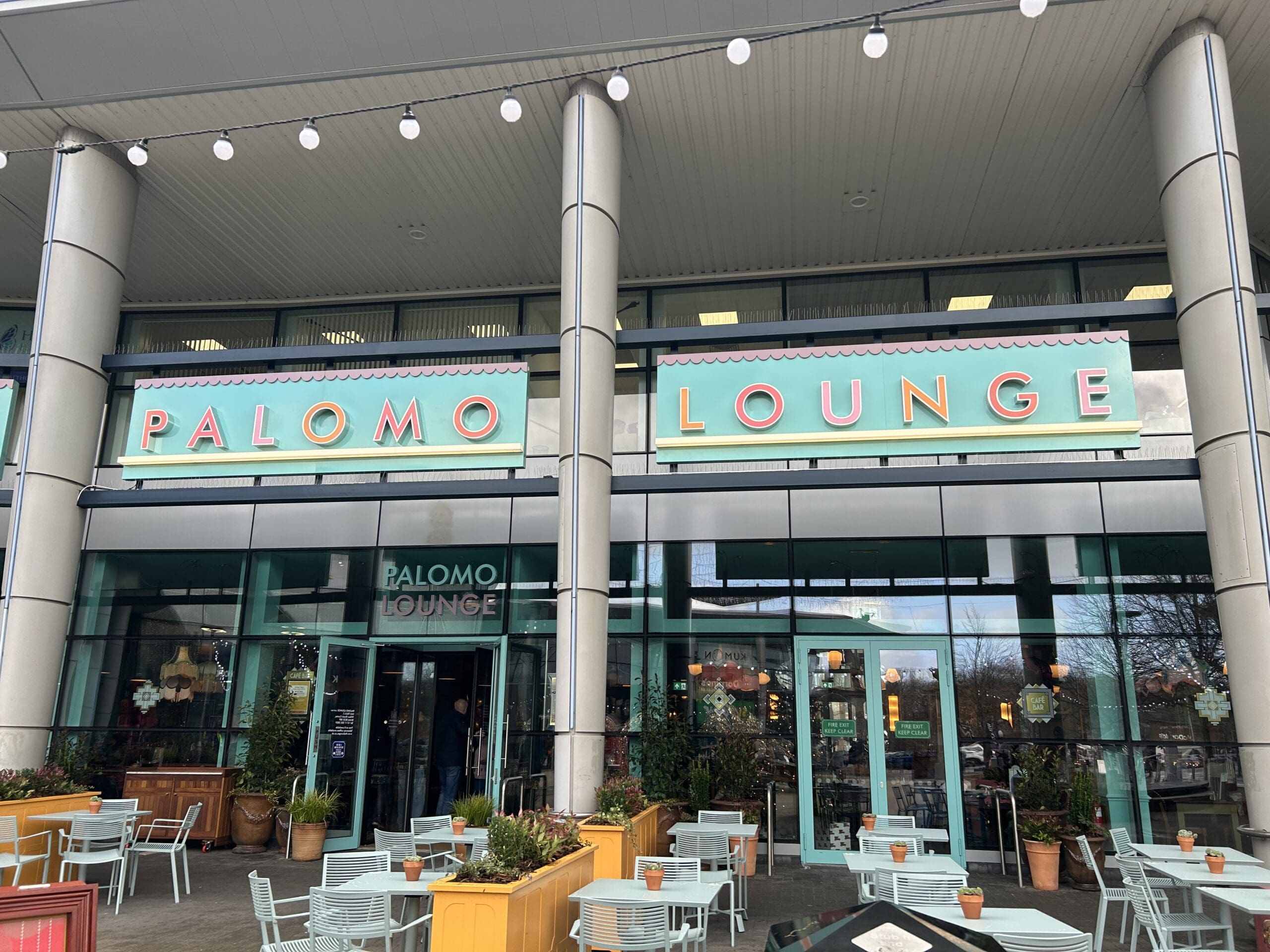 The exterior of Palomo Lounge, a vibrant café, features large glass windows, colorful signage, outdoor seating with pastel tables and chairs, and potted plants arranged around the entrance.