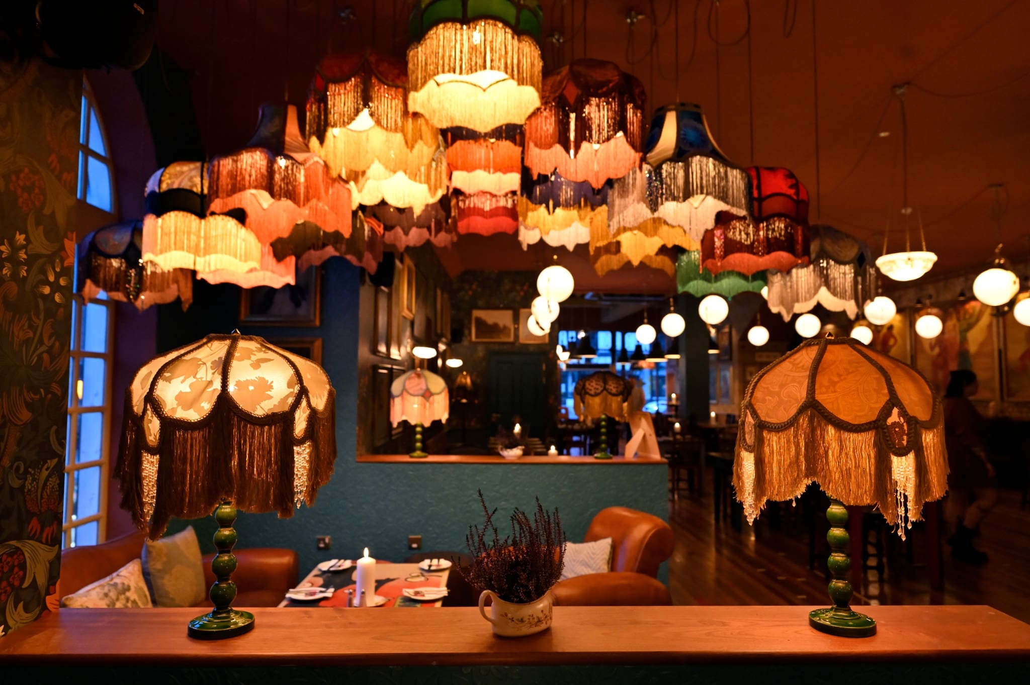 A cozy, warmly lit room sets the scene for our About Us story: vintage lampshades with fringes in various colors hang from the ceiling, creating an inviting, retro atmosphere. A teacup with flowers sits gracefully in the foreground.
