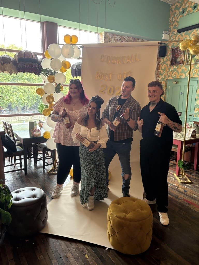 Four people stand smiling in front of a white and gold "Cocktail Host of 2023" backdrop, each holding a bottle. The scene is indoors with colorful decor, plants, and natural light from large windows.