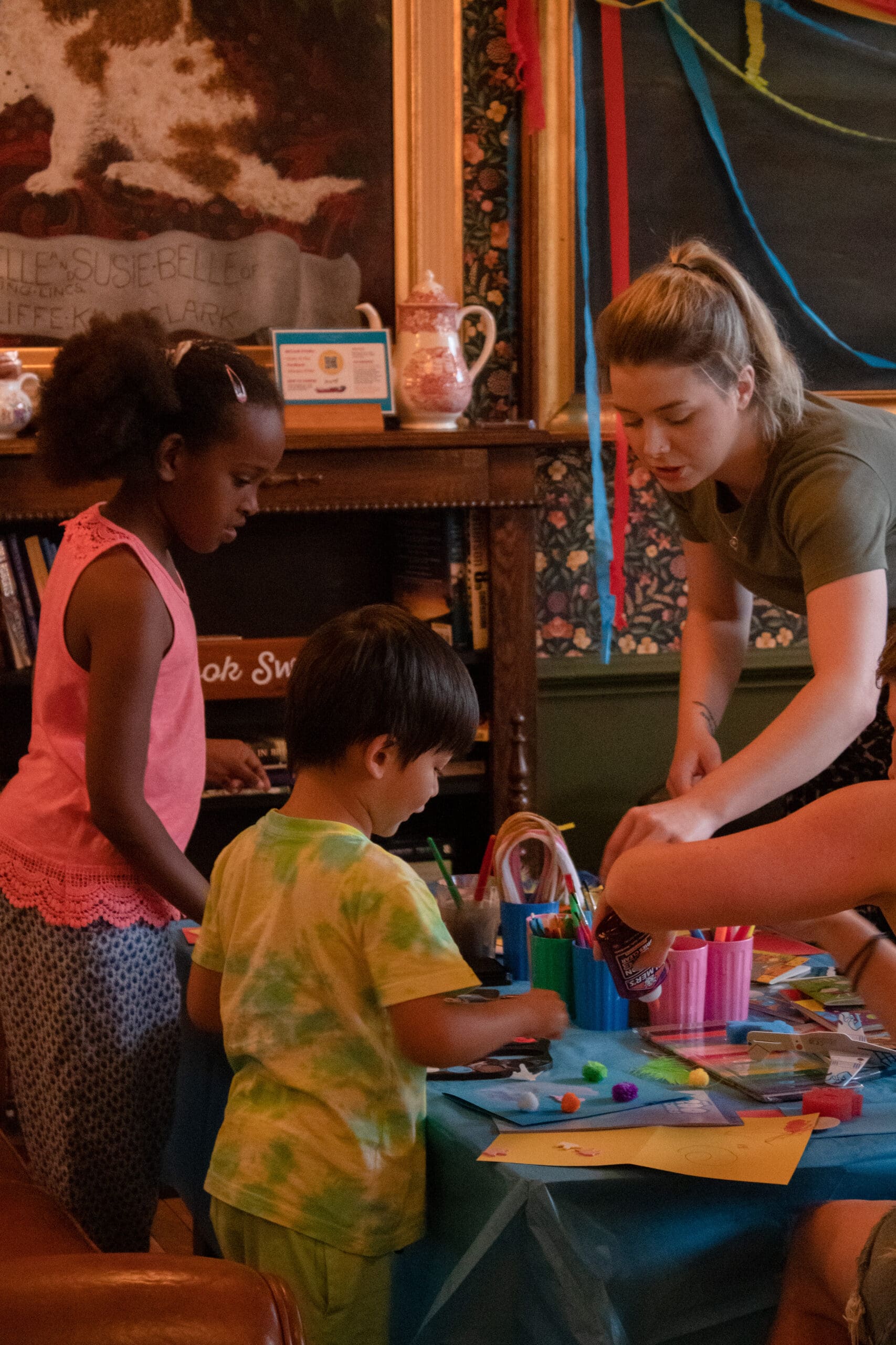 A young woman helps two children with arts and crafts at a colorful table, surrounded by art supplies in a cozy, decorated room. The children are focused on their creative projects.