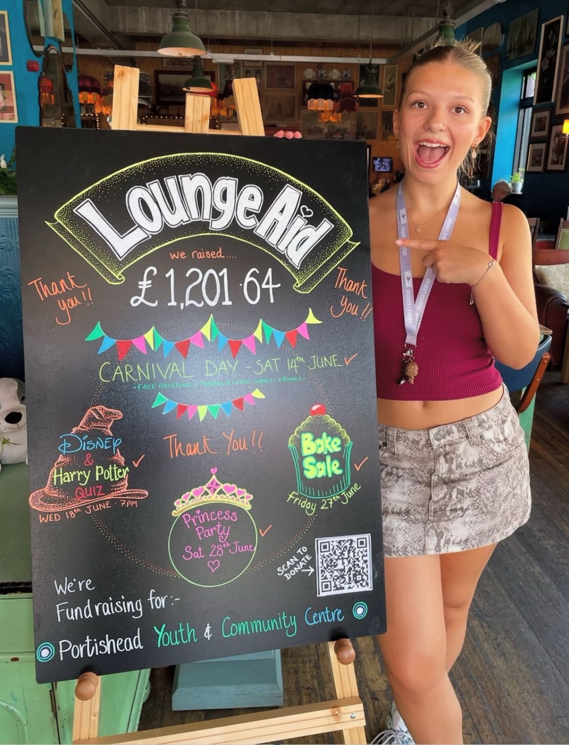A smiling young woman wearing a maroon tank top and patterned shorts points at a colorful chalkboard sign showing “LoungeAid” fundraising details and a total raised of £1,201.64, with event info and decorations.