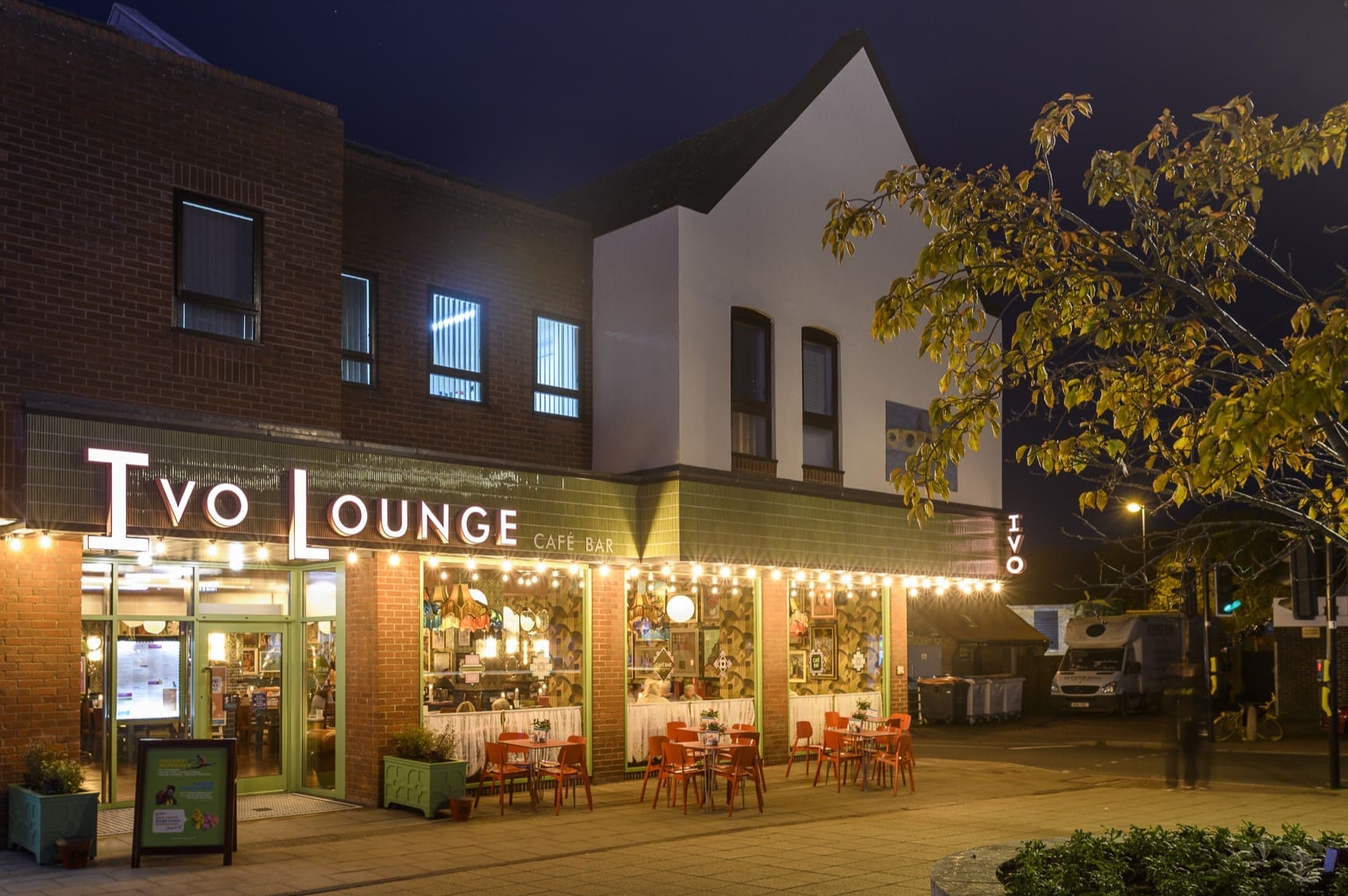 Ivo Lounge glows at night, its string lights shining on red chairs and tables outside. The two-story Ivo café bar sits beneath autumn leaves, while the quiet street sees only a few people passing by.