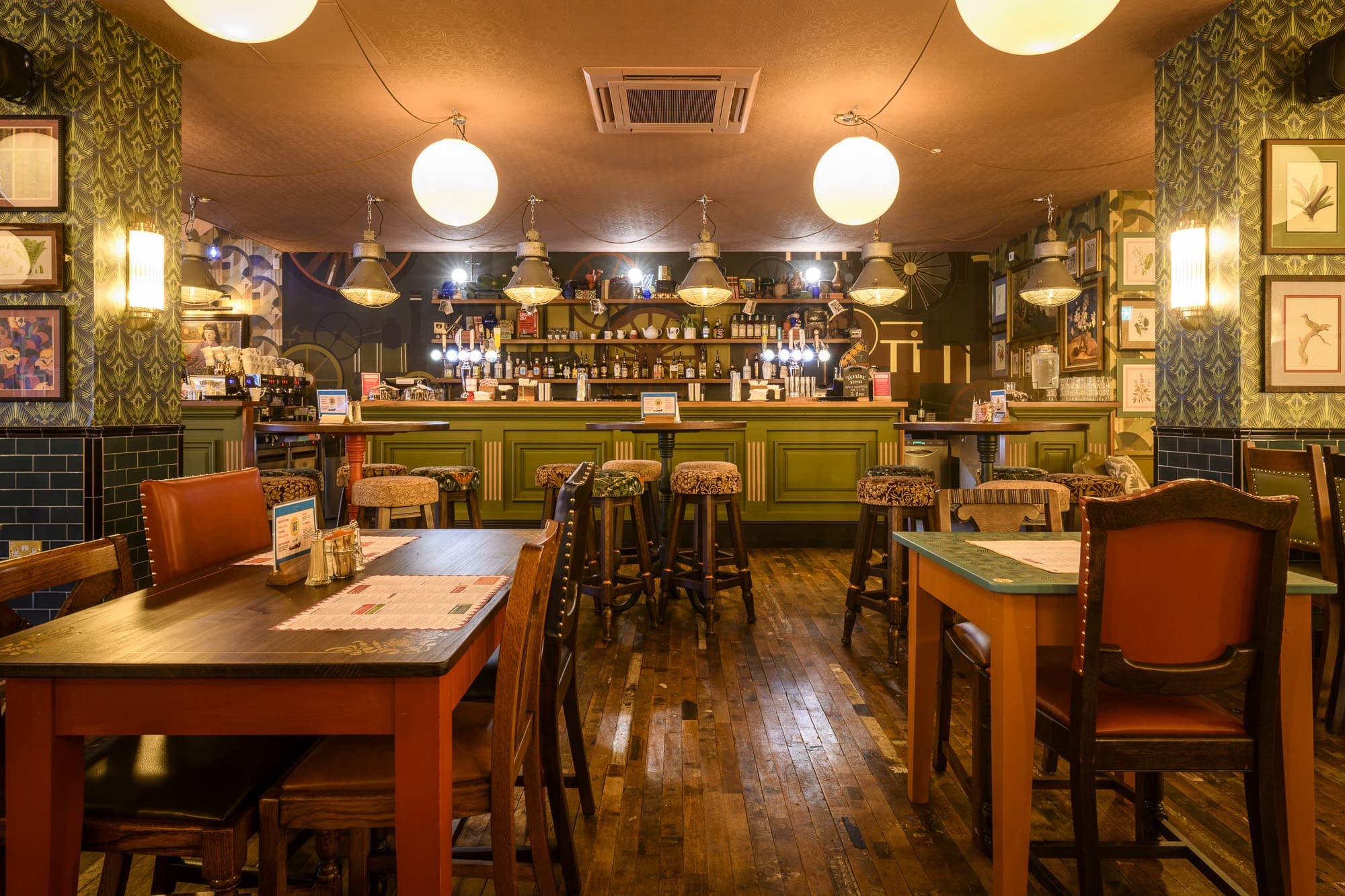 A warmly lit, cozy restaurant interior with wooden tables and chairs, hanging globe lights, framed pictures on the walls, and a well-stocked Ivo bar in the background. The space appears inviting and ready for customers.
