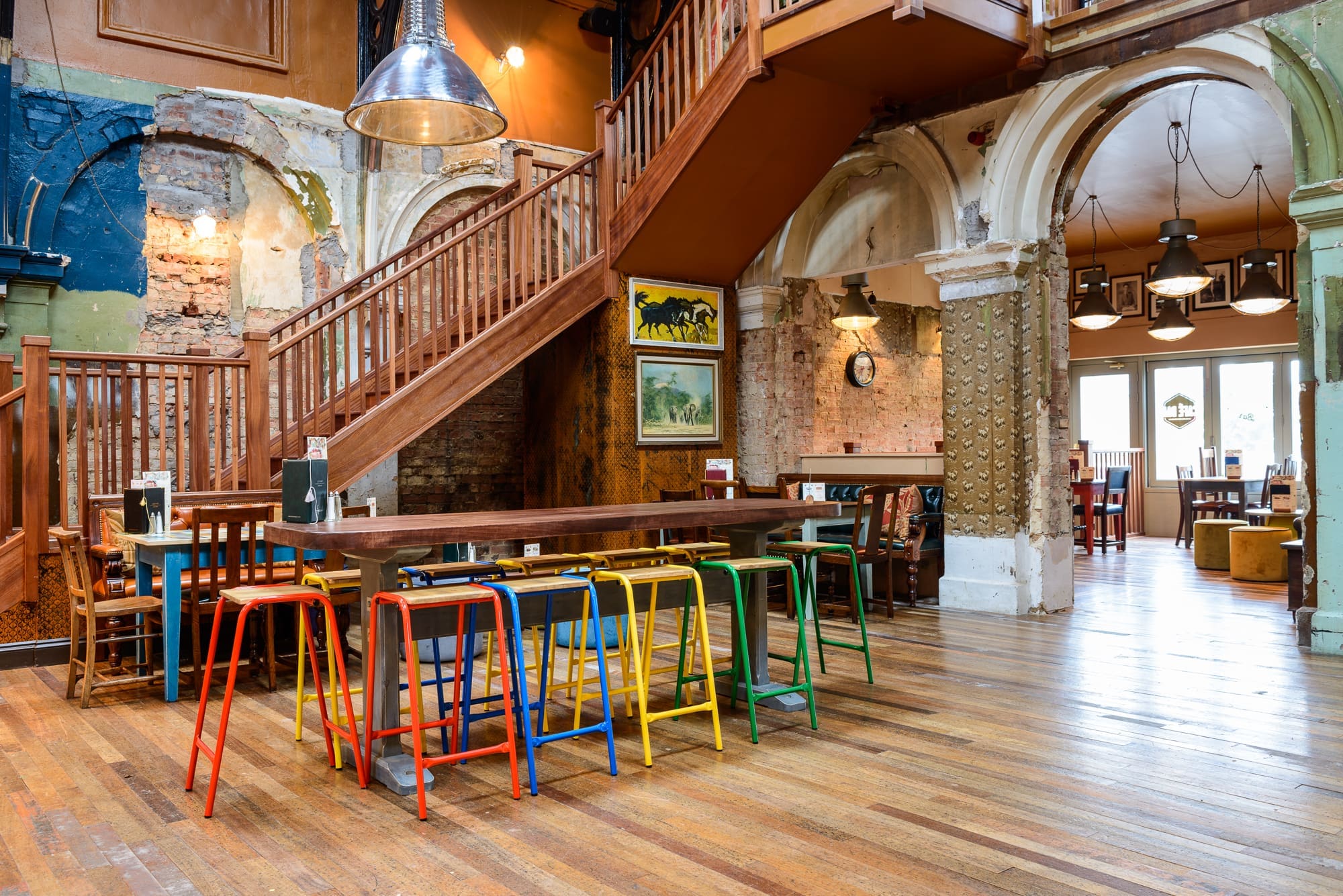 Spacious, rustic cafe interior with exposed brick walls, wooden floors, and a staircase. A long table is surrounded by colorful stools. Kino vibes fill the space as natural light streams through large windows in the background.