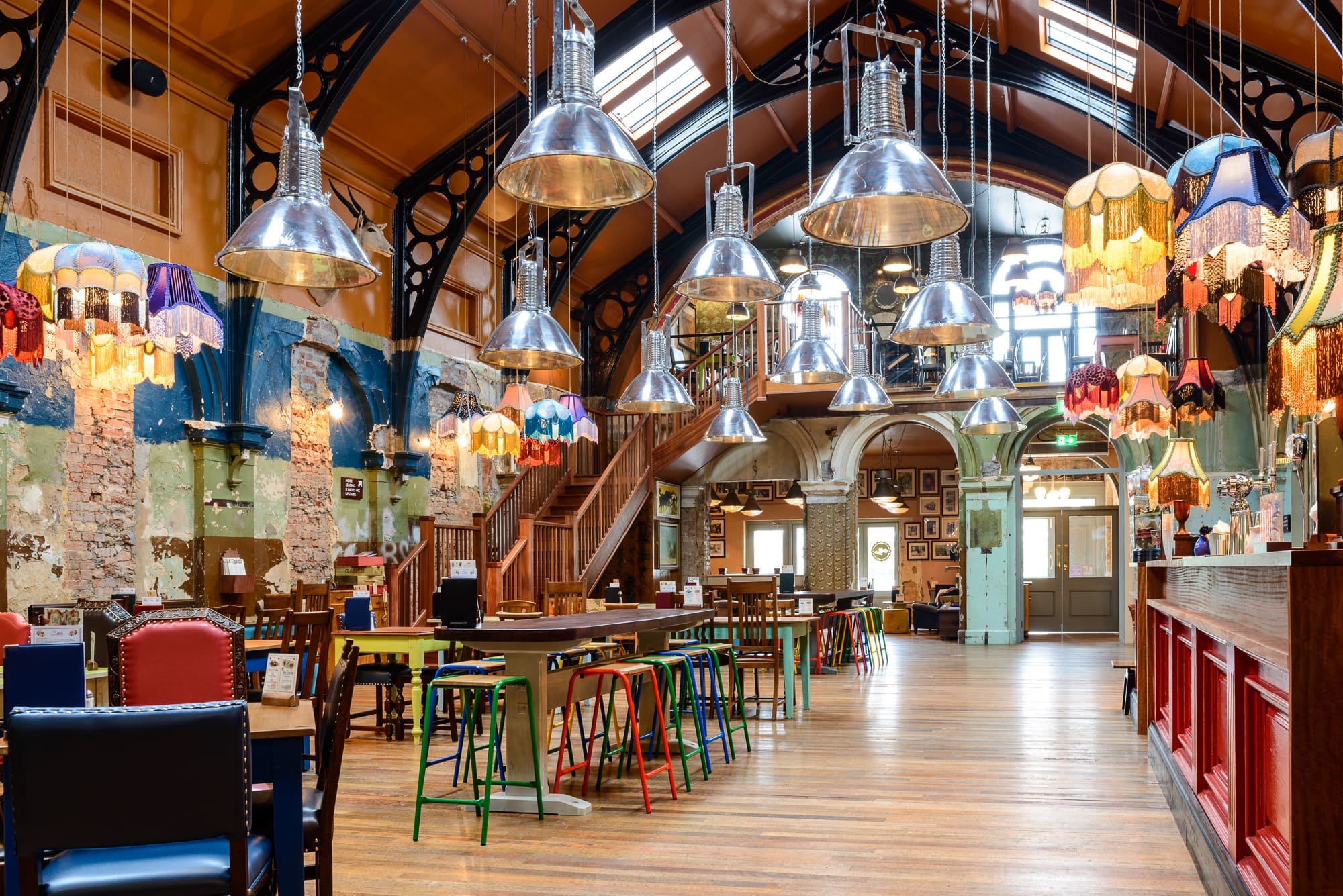 Bright, eclectic restaurant interior with colorful chairs and Kino-style stools, long wooden tables, exposed brick walls, and a mix of industrial and vintage hanging lamps under a high, skylit ceiling. A staircase leads to a mezzanine.