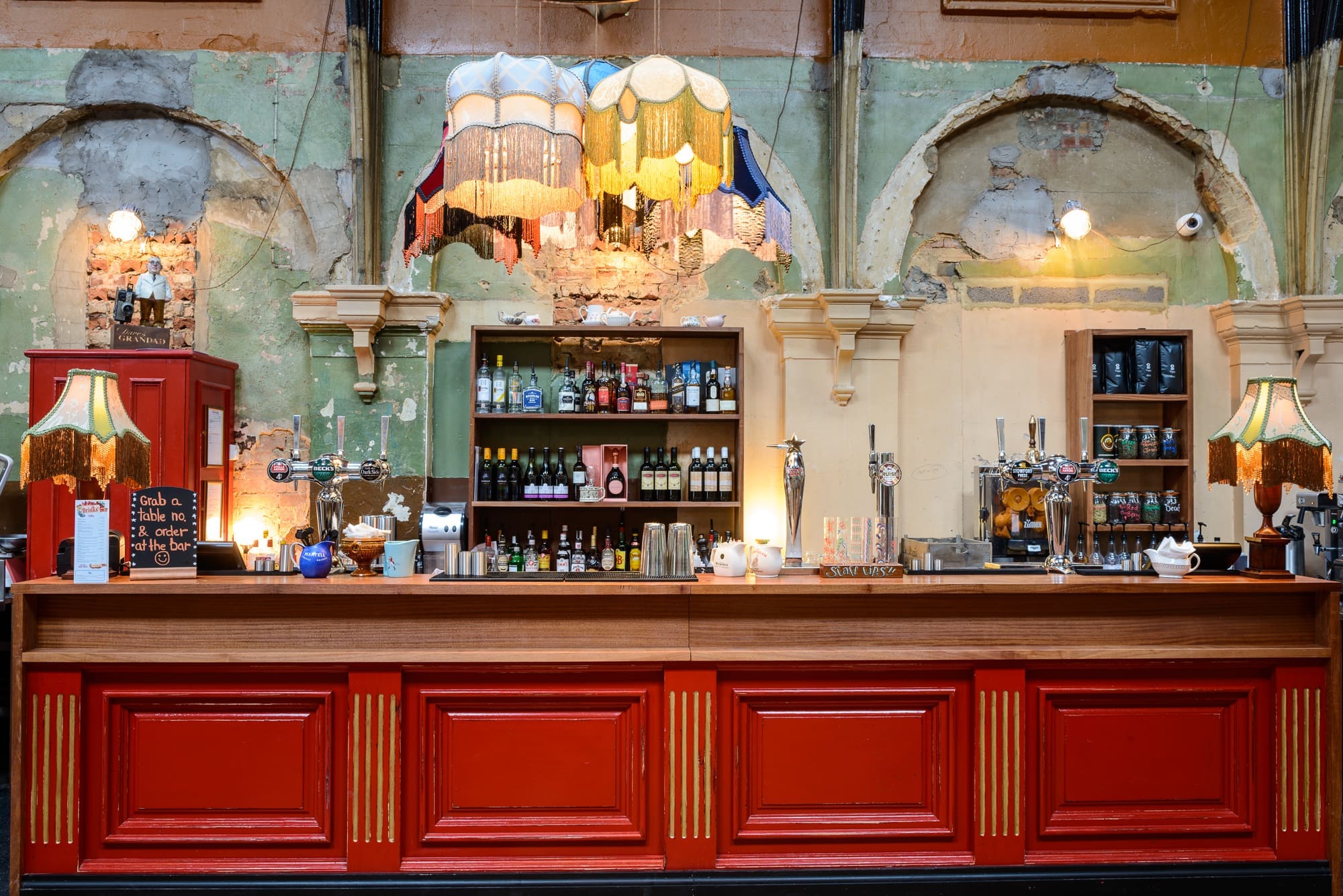 A vintage-style bar with a wooden counter, red panels, colorful hanging lamps, and Kino shelves stocked with bottles. The distressed walls, exposed brick, and arches create an eclectic, retro atmosphere reminiscent of an old-school cinema lounge.