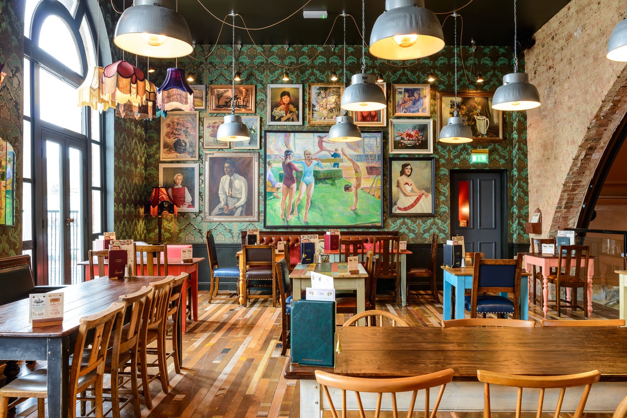 Eclectic restaurant interior with wooden tables and mismatched chairs, colorful Kino-inspired hanging lamps, and walls decorated with vintage paintings, including a large central artwork depicting people in a park. Natural light streams through tall arched windows.