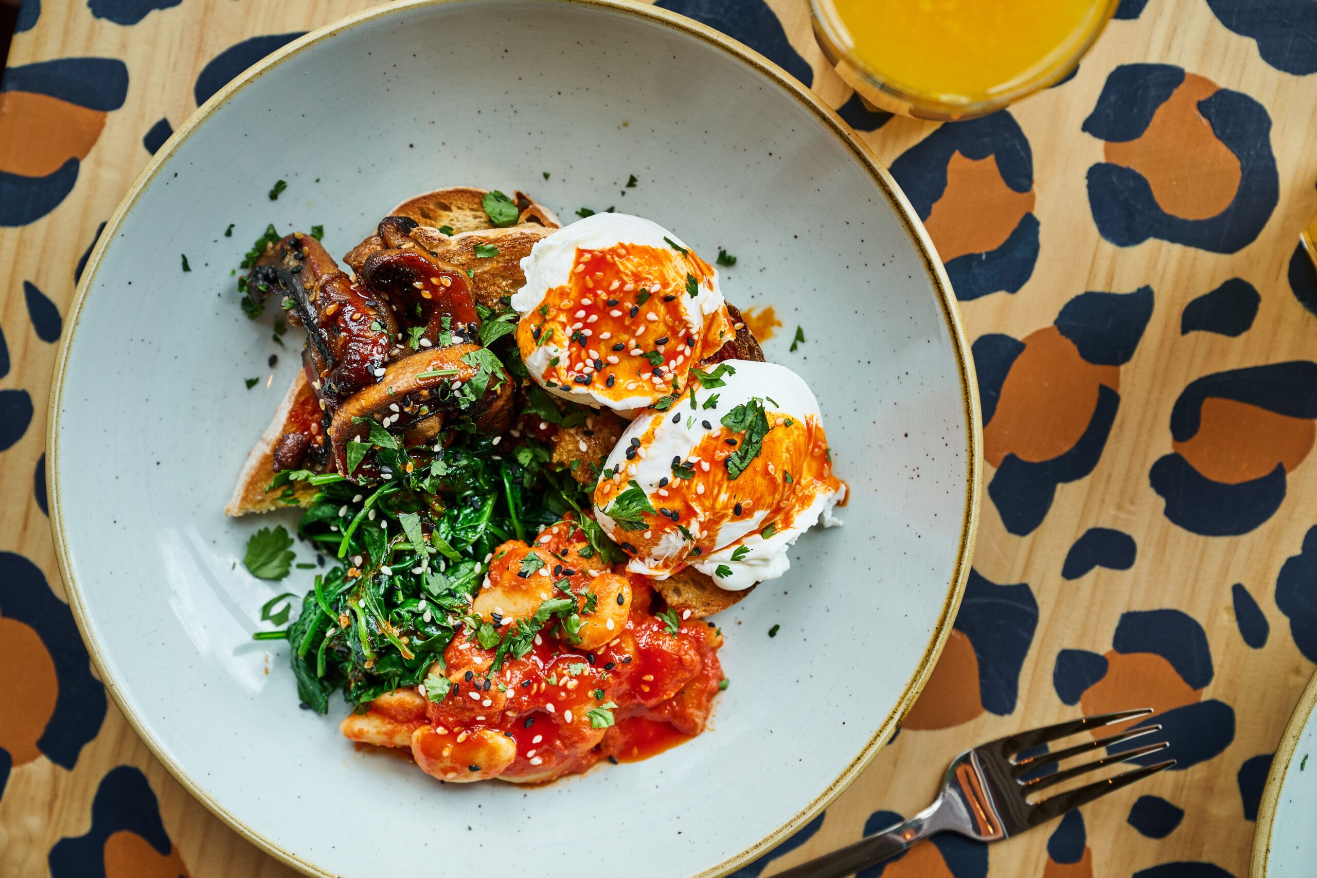 A plate with poached eggs topped with red sauce, sautéed spinach, mushrooms, and toast, garnished with herbs, sits on a table with a bold, abstract animal-print pattern. A fork and glass are nearby.