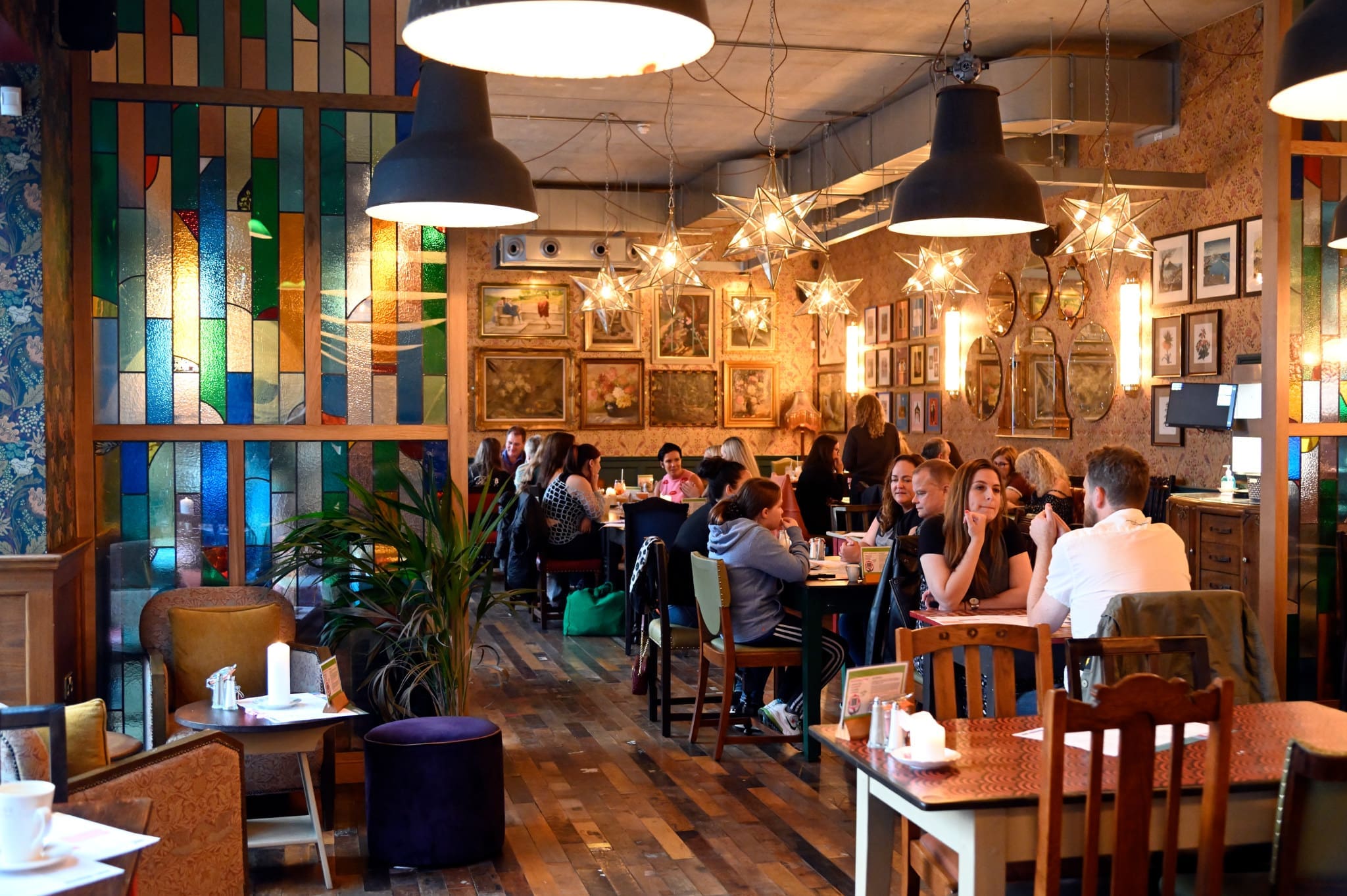 A cozy Liberto restaurant with colorful stained glass, hanging lights, and framed pictures on the walls. Groups of people are dining and chatting at wooden tables in a warm, inviting atmosphere.