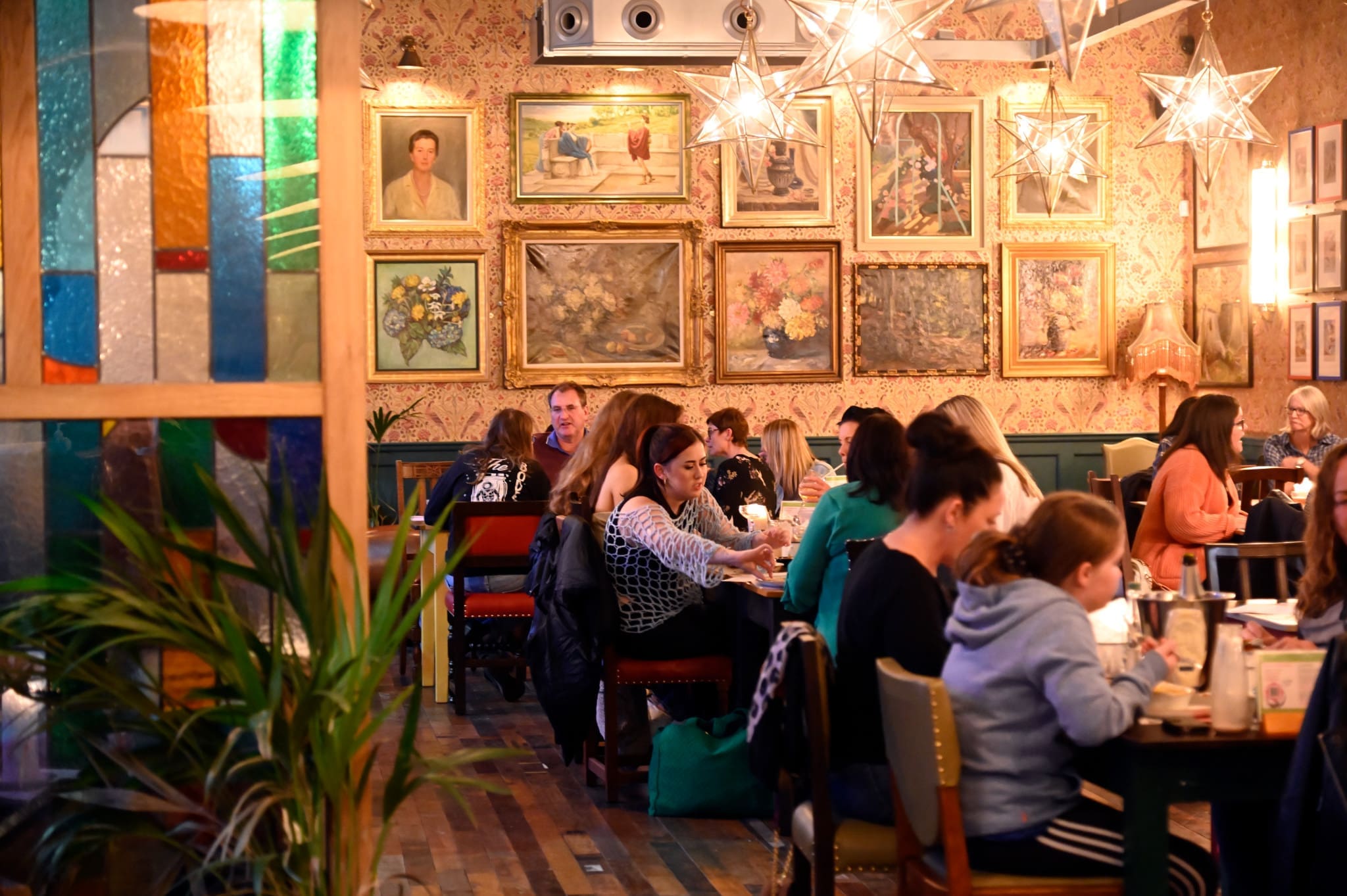 People sit at tables in Liberto, a cozy restaurant with warm lighting, colorful stained glass, and framed artwork on the walls. The lively atmosphere encourages diners to engage in conversation and savor their meals.