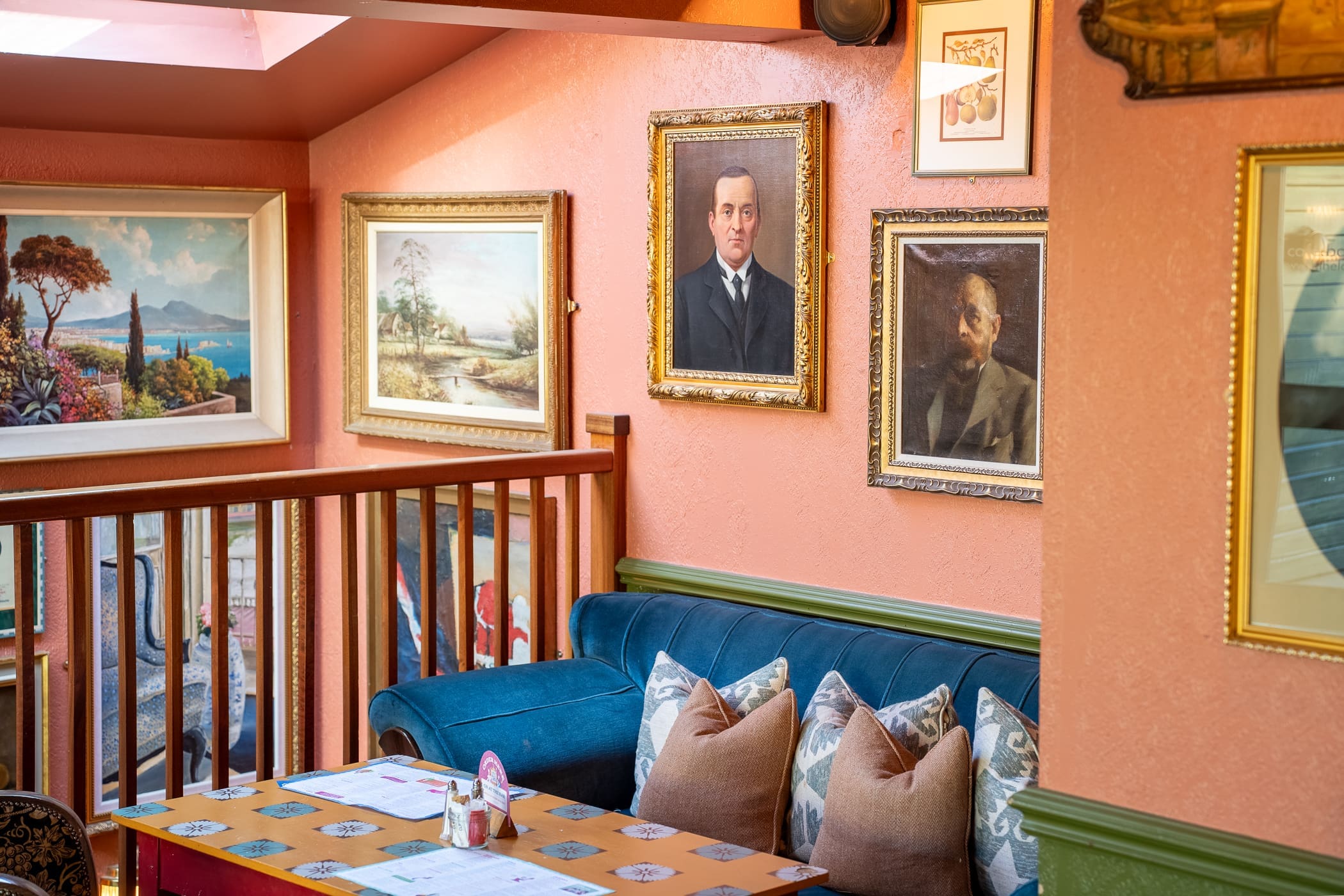 A cozy Loco cafe corner with a blue velvet sofa, beige pillows, a wooden table with menus, and framed paintings and portraits hanging on peach-colored walls above a wooden railing.