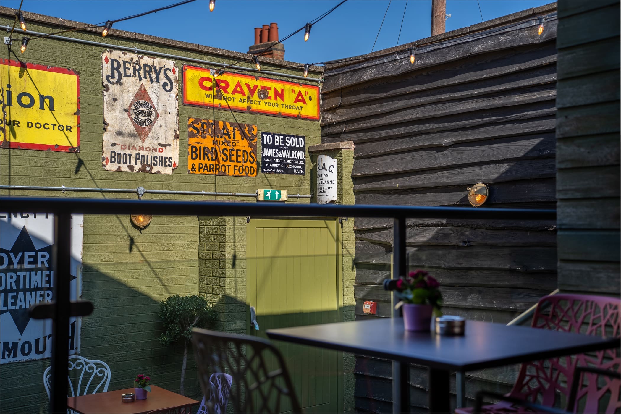 A sunny outdoor café area with string lights, colorful vintage advertisement signs on the walls, potted plants on tables, and a mix of pink and white Loco metal chairs and tables.