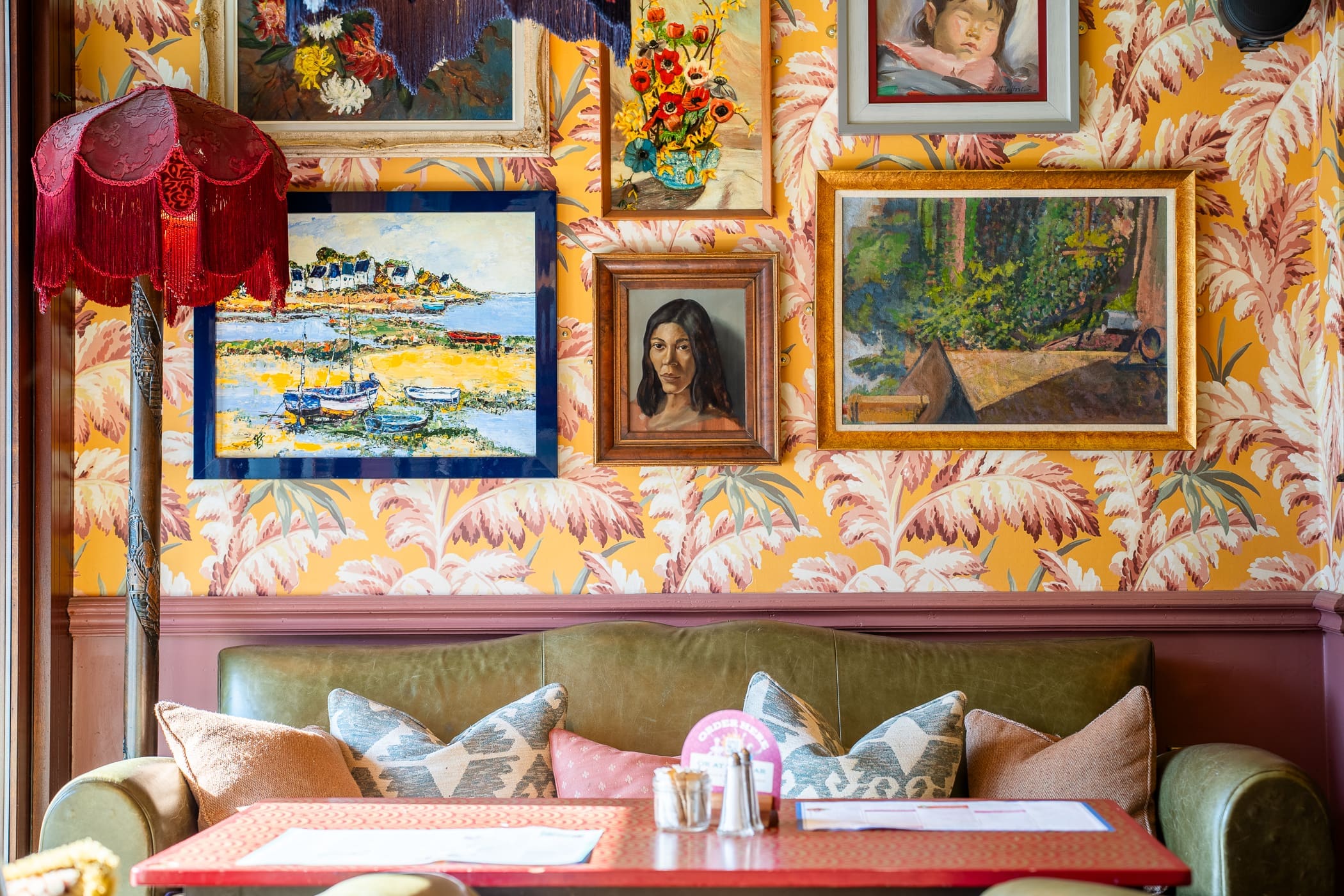 A cozy cafe seating area with a green sofa, colorful cushions, a red table, and a wall decorated with various framed paintings and a red vintage lampshade. The bold floral wallpaper adds a touch of Loco charm to the space.