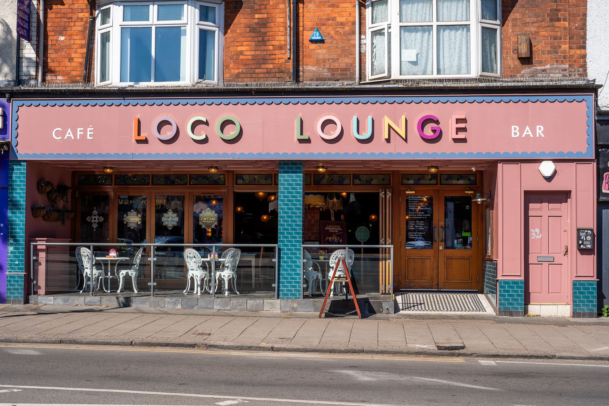 Colorful café and bar called "Loco Lounge" features playful, multicolored letters above large windows, outdoor seating with white chairs and tables, and teal-tiled columns on a lively city street.