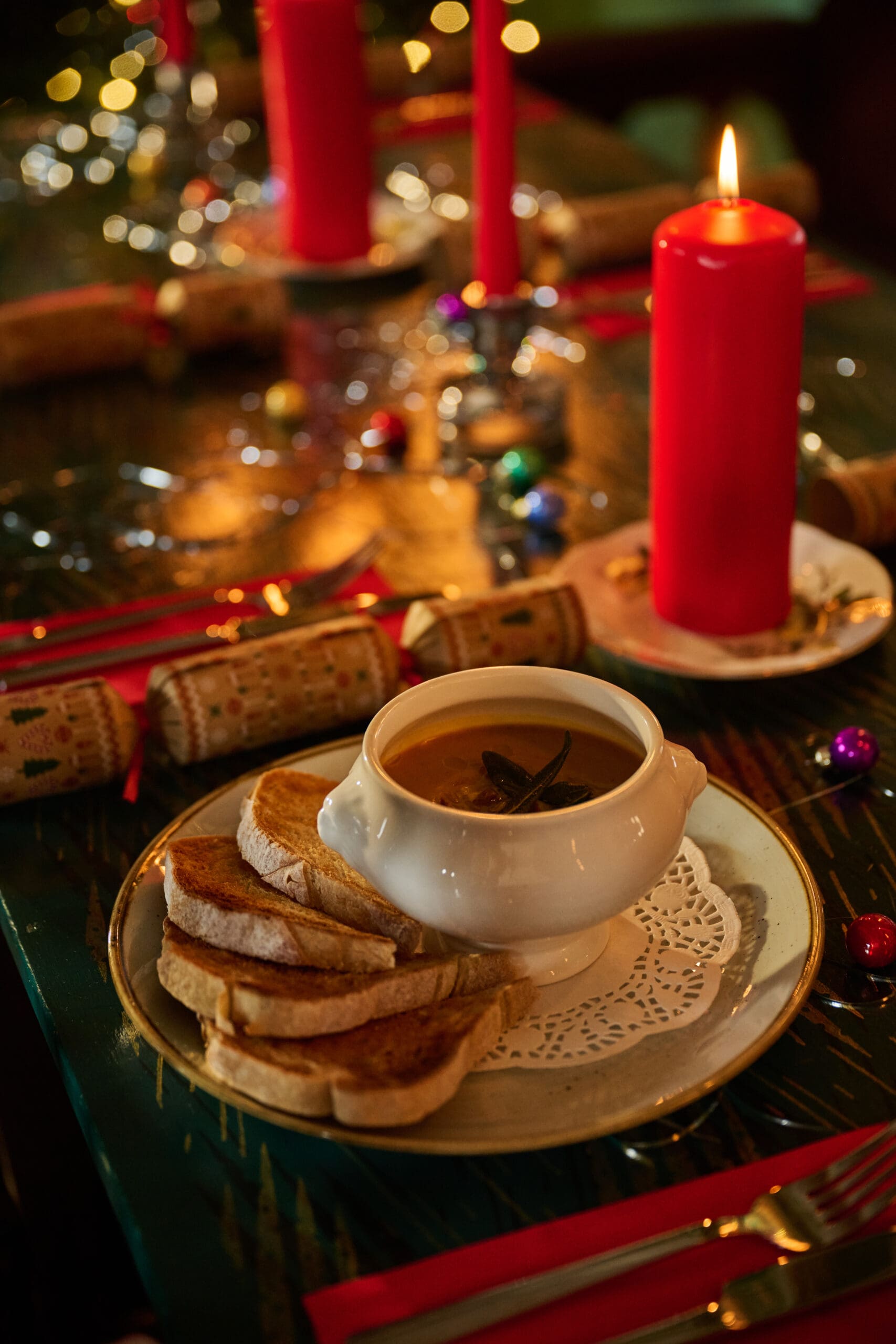 A festive table setting with a bowl of soup and four slices of toasted bread on a plate, surrounded by red candles, Christmas crackers, and holiday decorations.