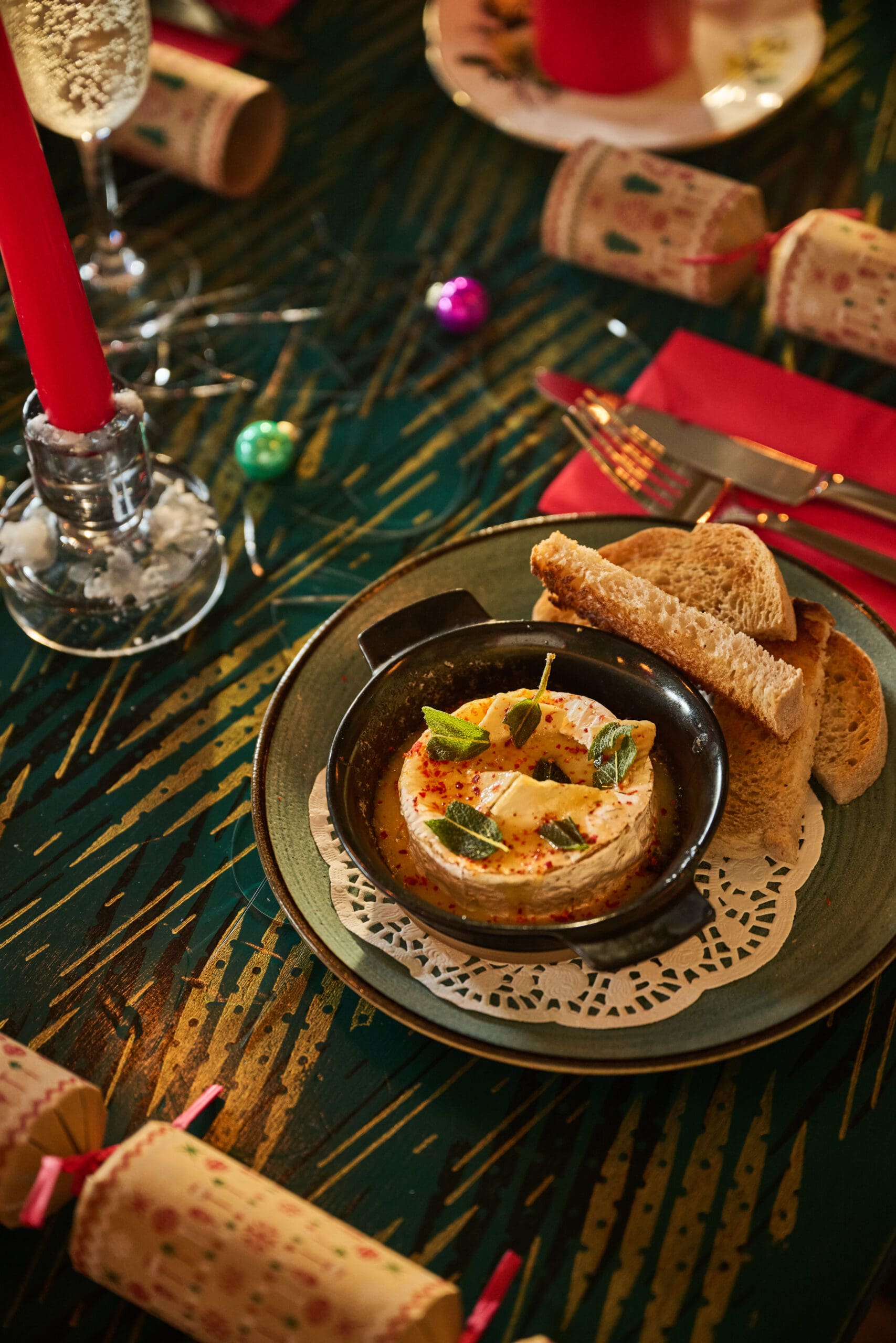 A plate with baked cheese topped with herbs and spices, served with sliced bread, sits on a festive table set with red napkins, crackers, candles, and holiday decorations.