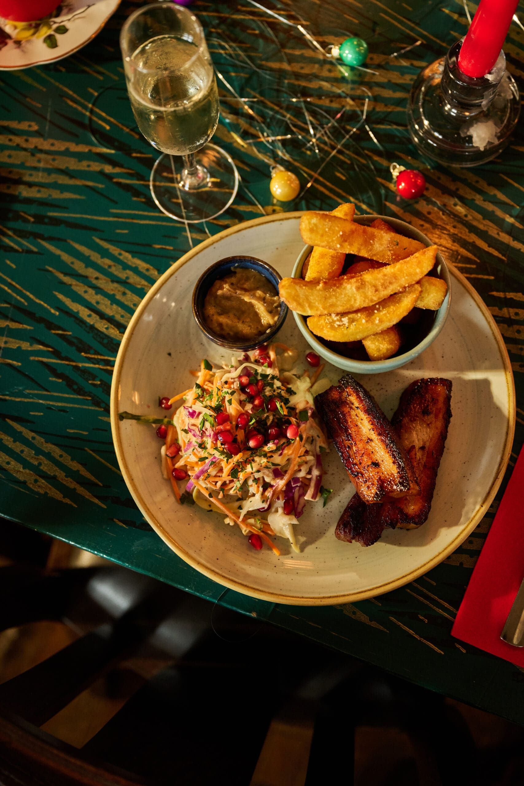 A plate with thick-cut fries, grilled meat slices, creamy dipping sauce, and a fresh salad topped with pomegranate seeds, next to a glass of white wine on a green patterned table with holiday decorations.