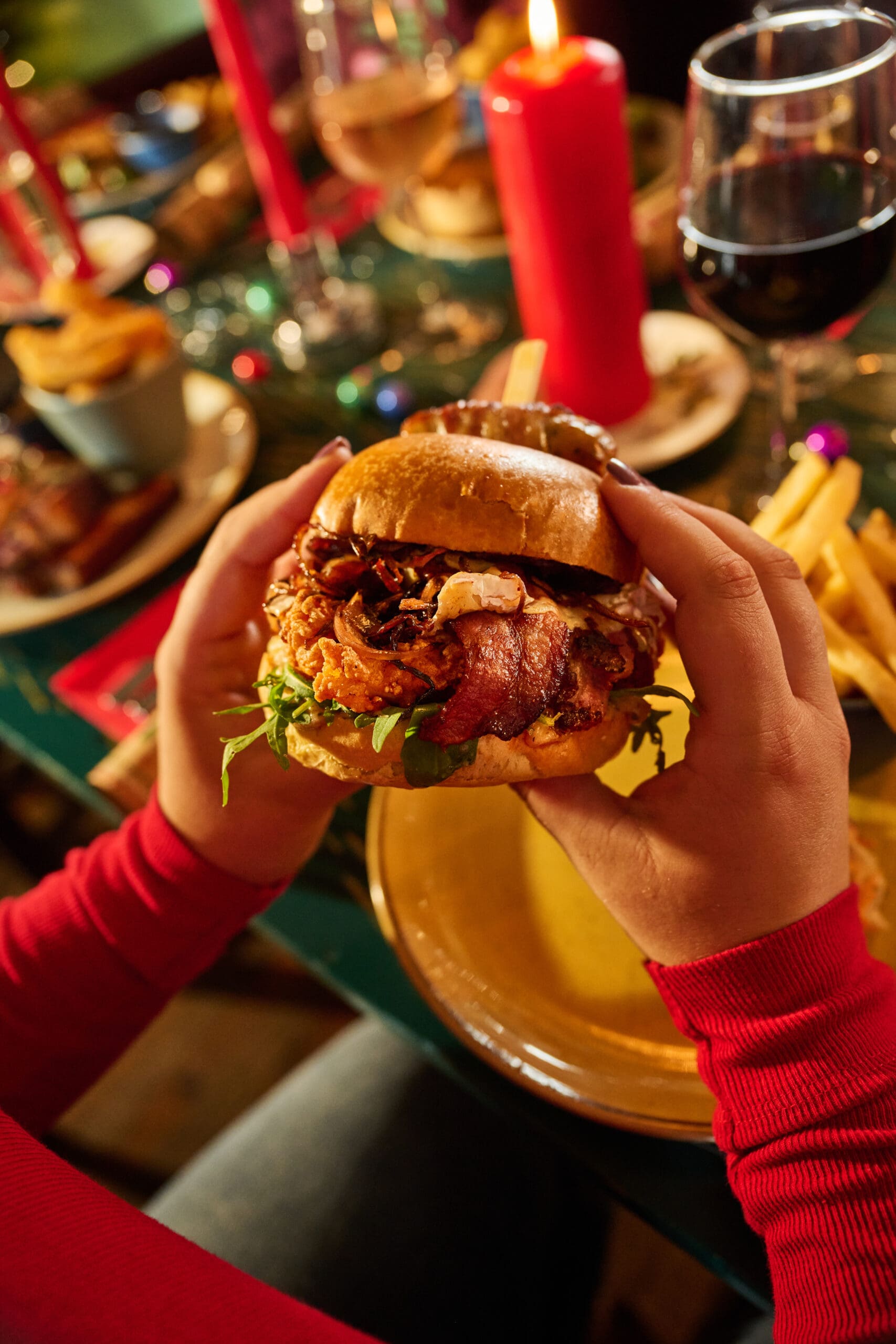 A person in a red sweater holds a large gourmet burger with lettuce, bacon, and crispy onions. In the background, there are fries, wine glasses, and lit red candles on a festive dinner table.