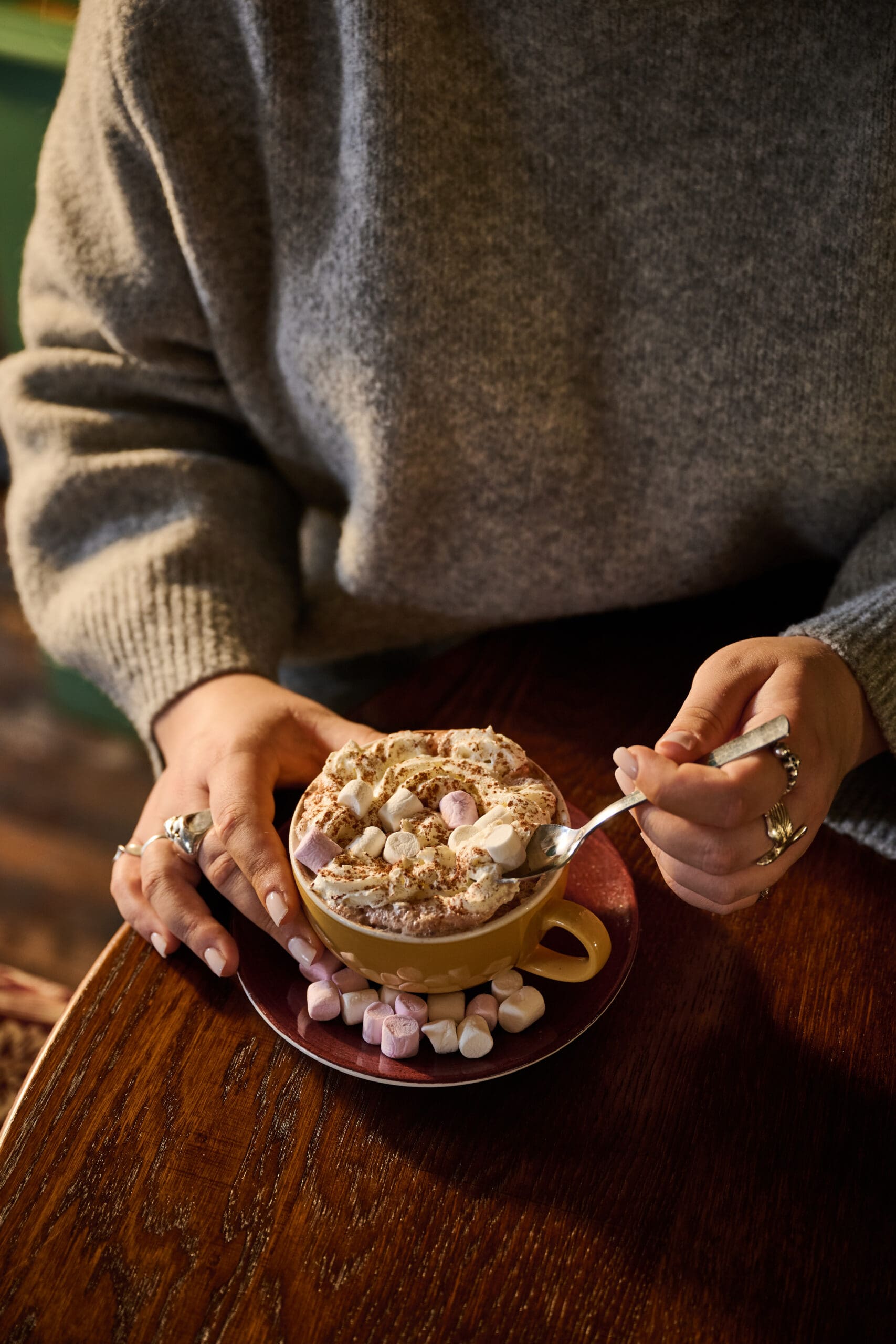 A person in a gray sweater sits at a wooden table, holding a spoon over a mug of hot chocolate topped with marshmallows and cocoa, with extra marshmallows scattered on the saucer.