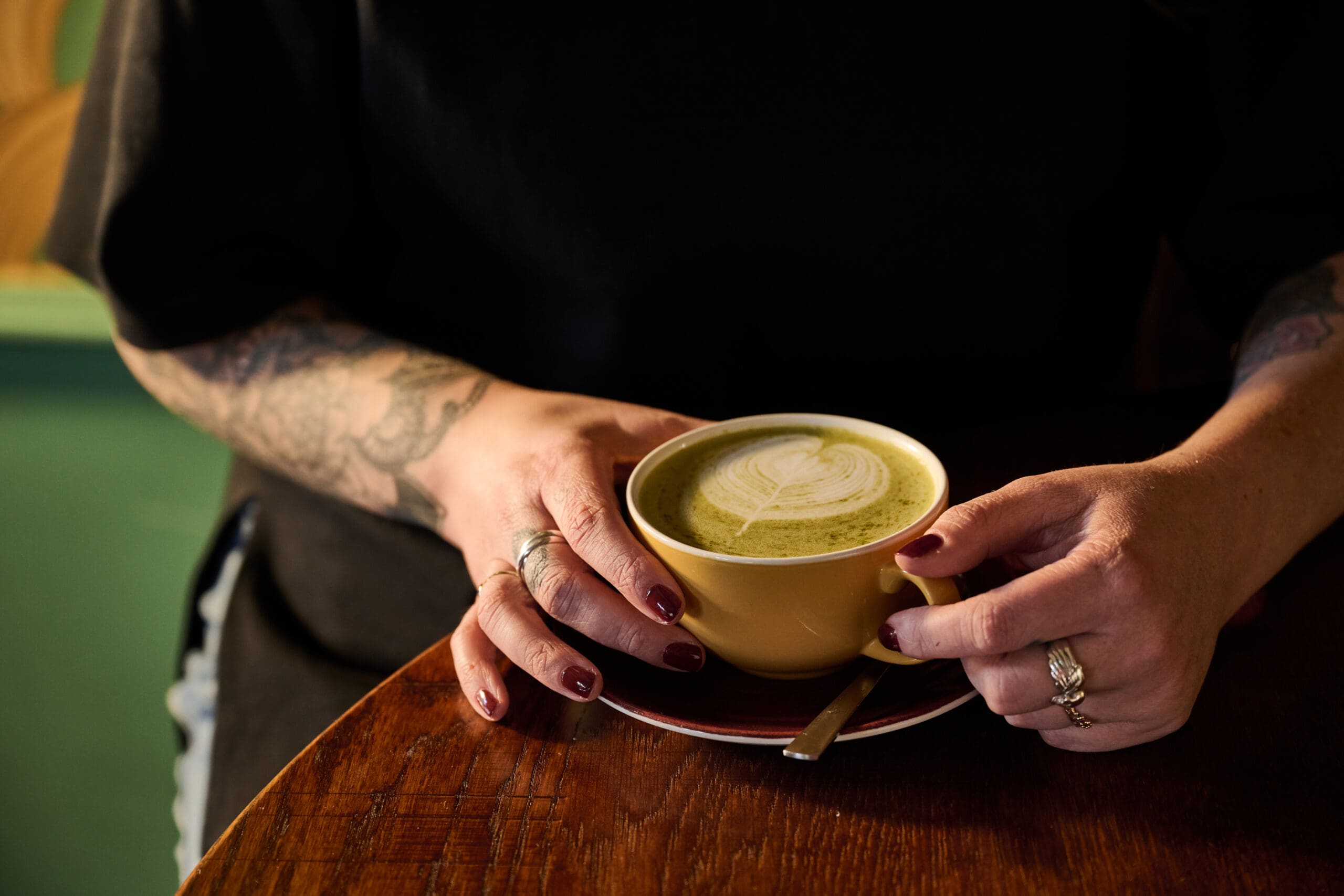 A person with tattoos and dark nail polish holds a yellow cup of latte with latte art on top, resting on a wooden table with a small spoon on the saucer.