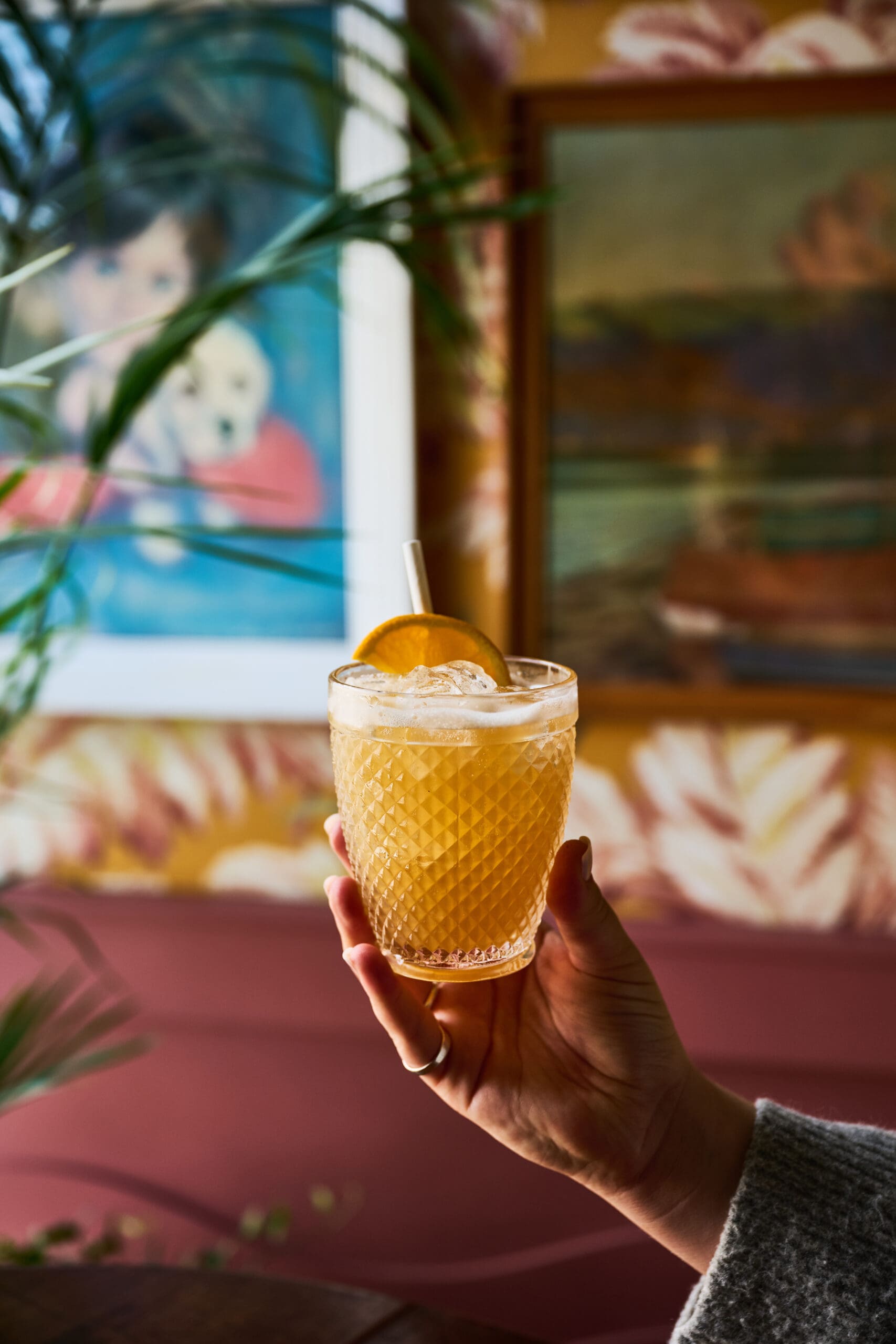 A hand holds a textured glass filled with an orange cocktail, topped with ice and garnished with an orange slice and a straw. Colorful, blurred artwork and leafy plants are visible in the background.