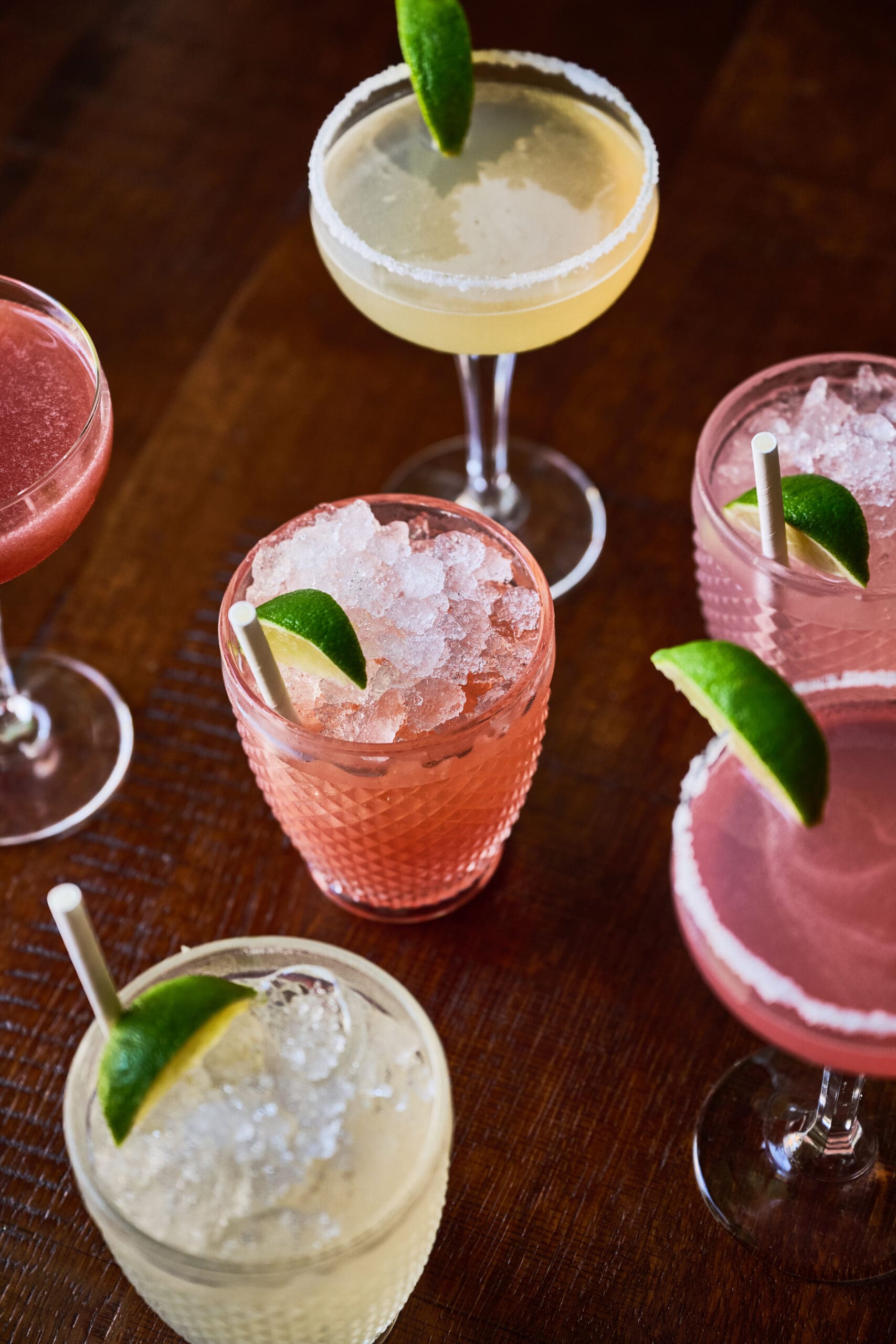 An overhead view of six colorful cocktails in various glasses, some with crushed ice and lime wedges, garnished with salted rims, set on a dark wooden surface.