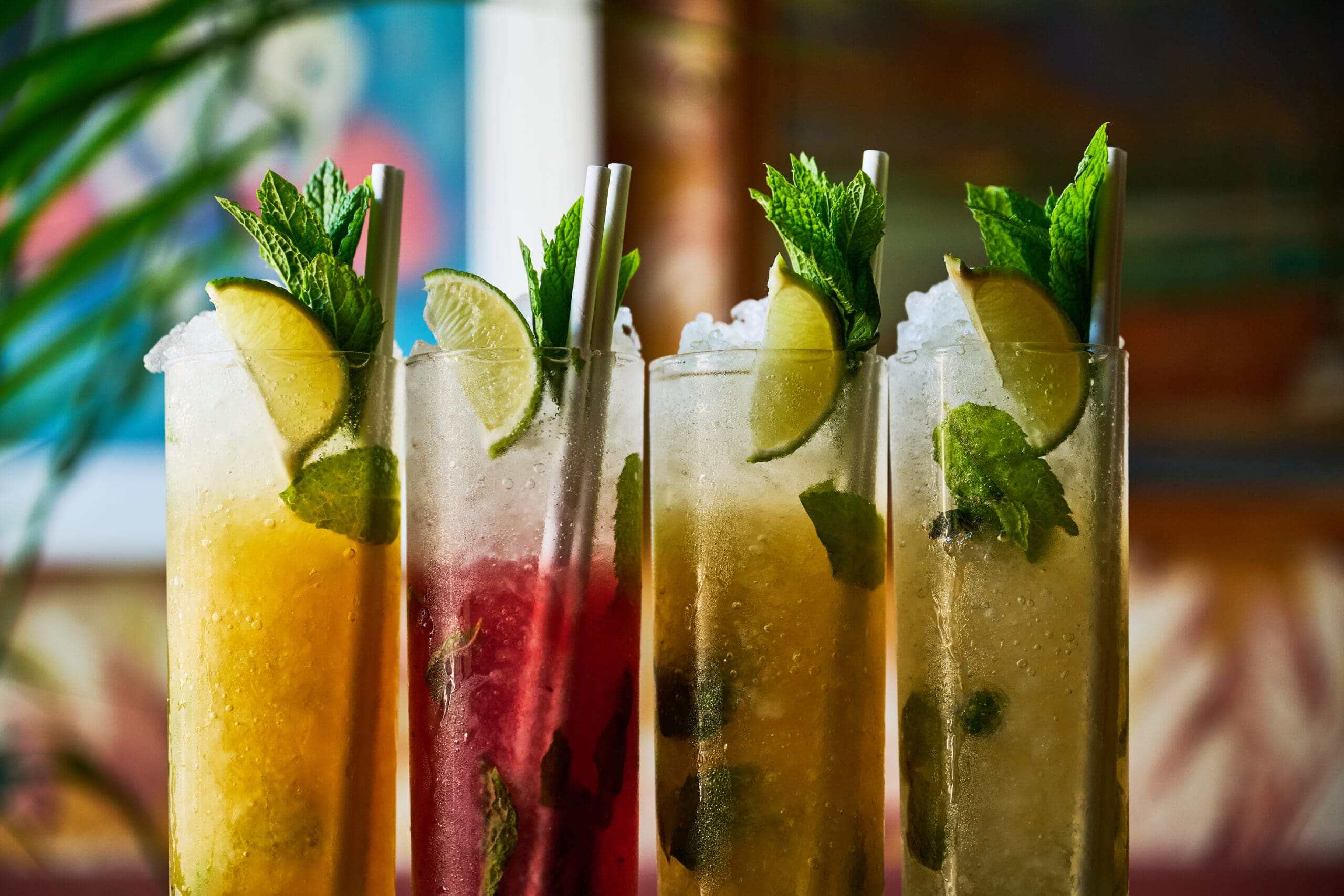 Four tall glasses of colorful, iced cocktails garnished with fresh mint sprigs and lime wedges, each with a straw, set against a blurred, vibrant background.