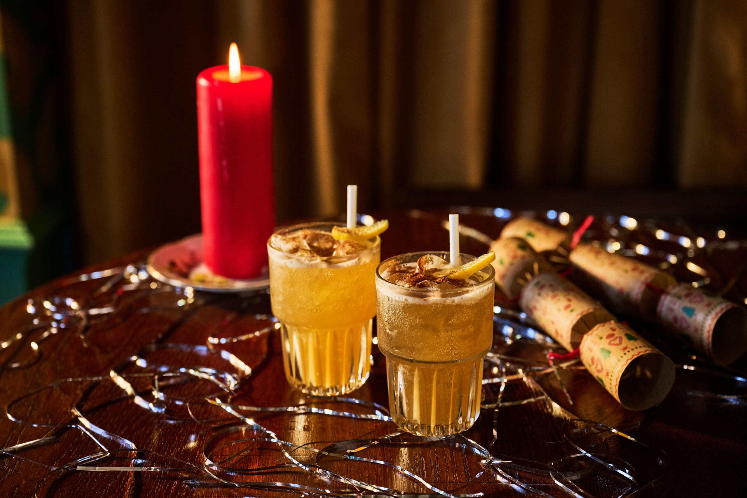 Two cocktails with lemon twists on a wooden table, surrounded by string lights. A red candle and two decorative Christmas crackers are also on the table, creating a festive atmosphere.