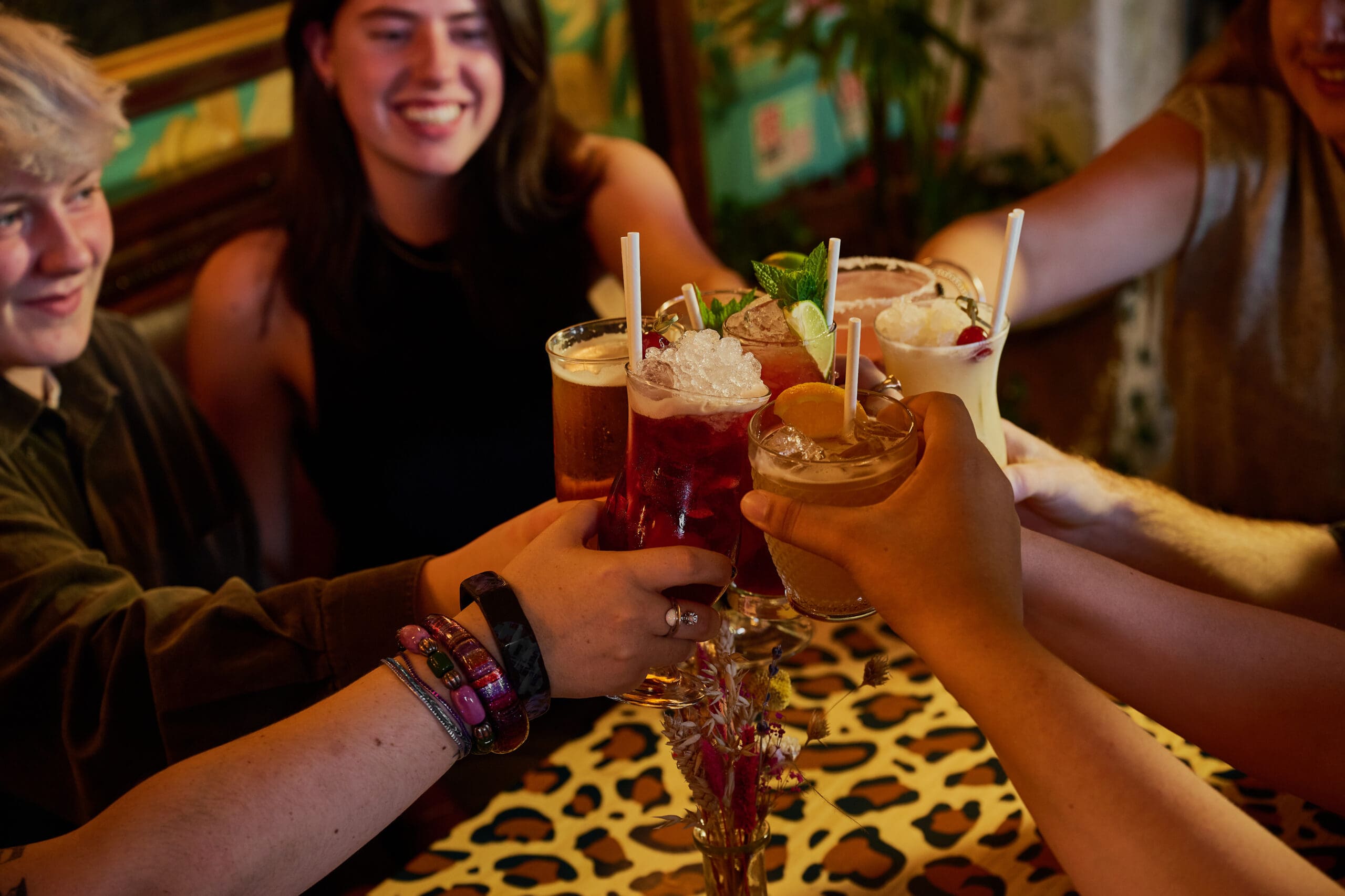 A group of friends sit around a table with a leopard print tablecloth, raising various colorful cocktails in a toast, smiling and enjoying each other's company in a cozy, warmly lit setting.