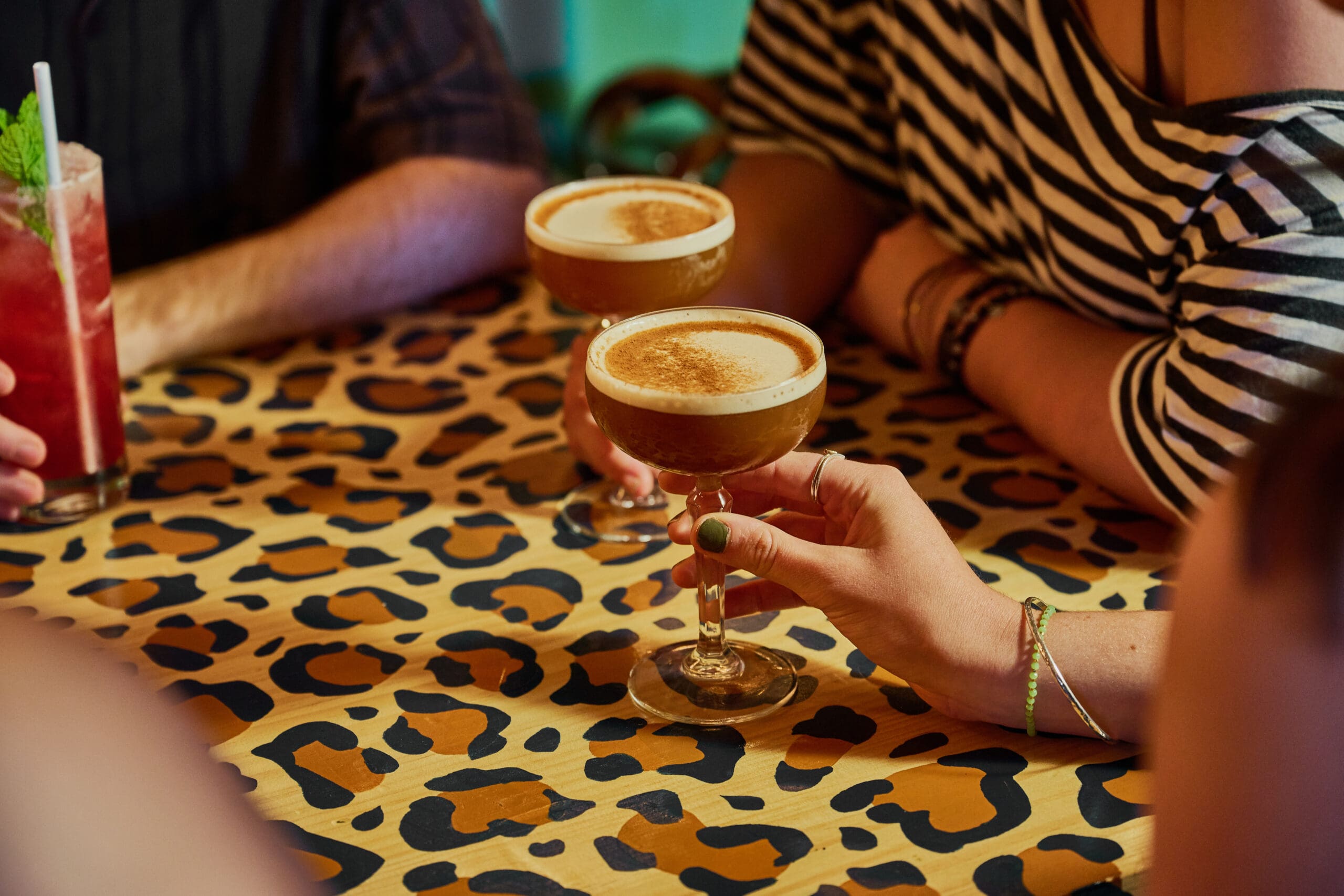 People sit around a leopard-print table, holding cocktail glasses filled with frothy drinks. One person wears a striped shirt and holds a drink; another has a minty red cocktail with a straw.