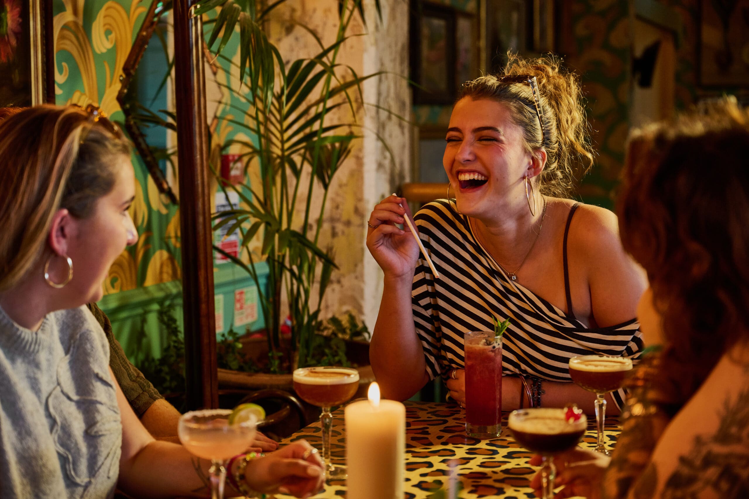 Three women sit at a table with cocktails, laughing and enjoying themselves in a warmly lit, colorful lounge bar adorned with lively decor and a glowing candle at the center.