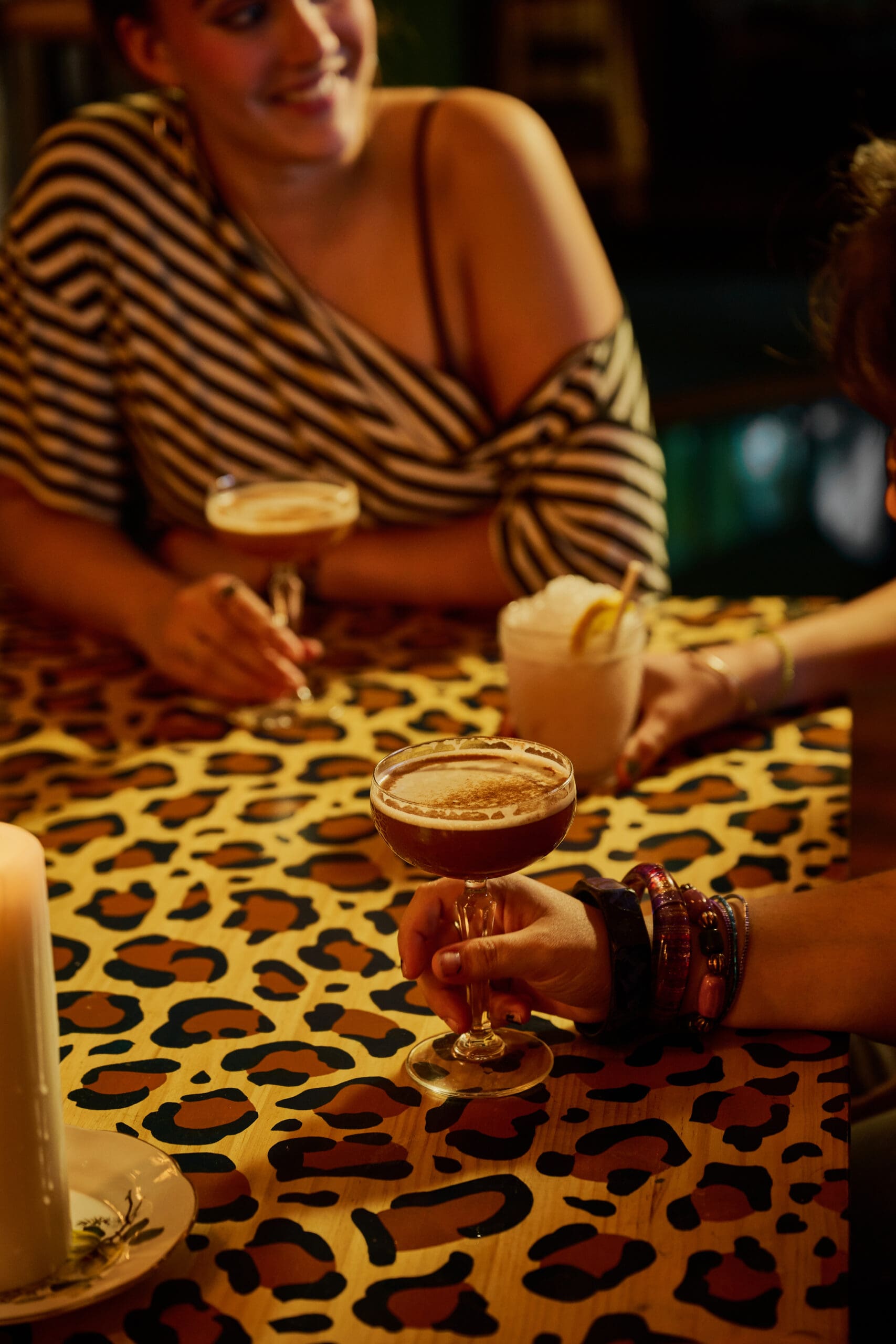 Two people sit at a table with a leopard print surface, holding cocktails and chatting. One person wears a striped off-the-shoulder top, and the scene is warmly lit, creating a cozy, relaxed atmosphere.