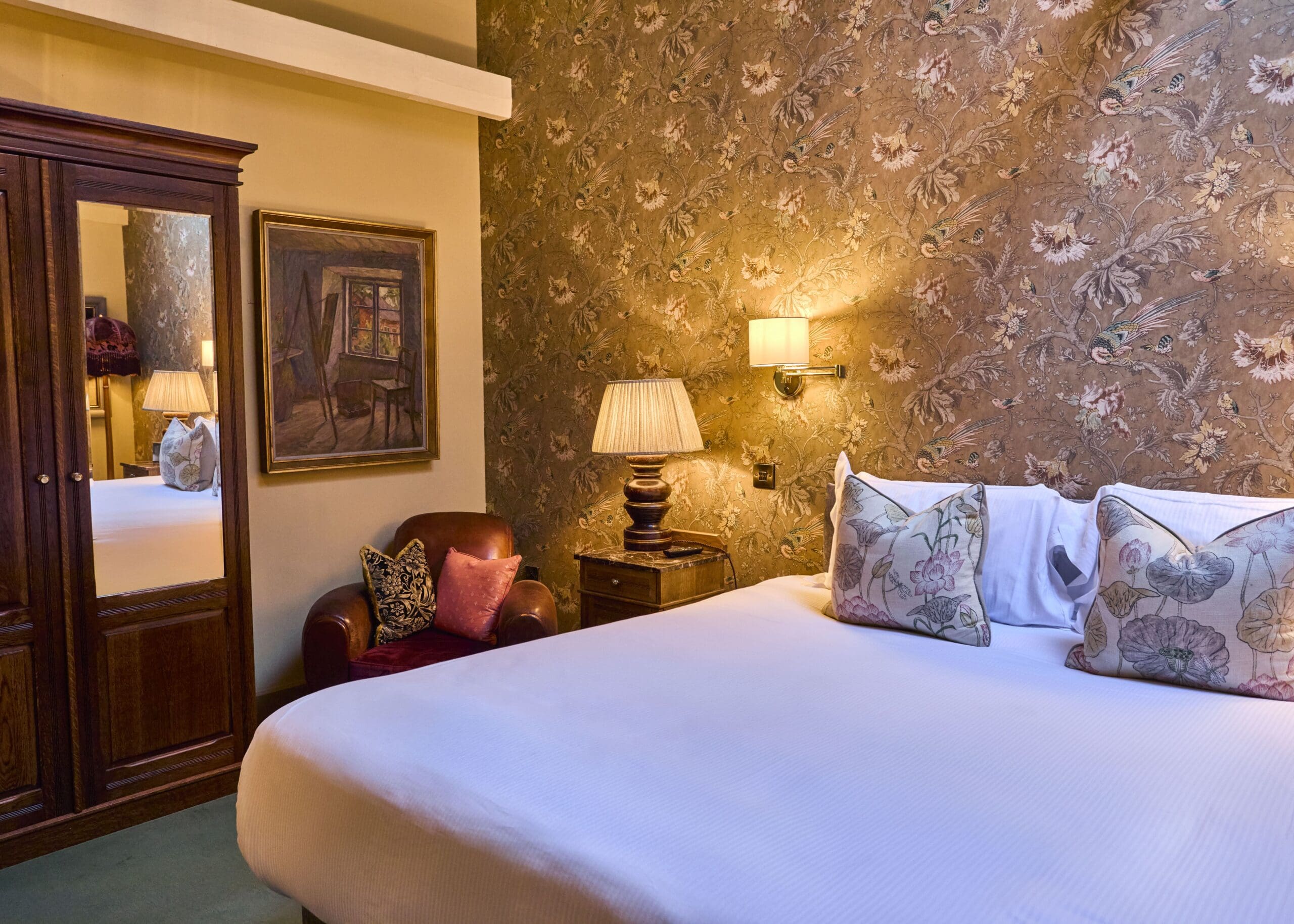 A cozy hotel room with a large bed, floral wallpaper, decorative pillows, a leather armchair, a lamp on a wooden nightstand, a wardrobe with mirrored doors, and a framed painting on the wall.