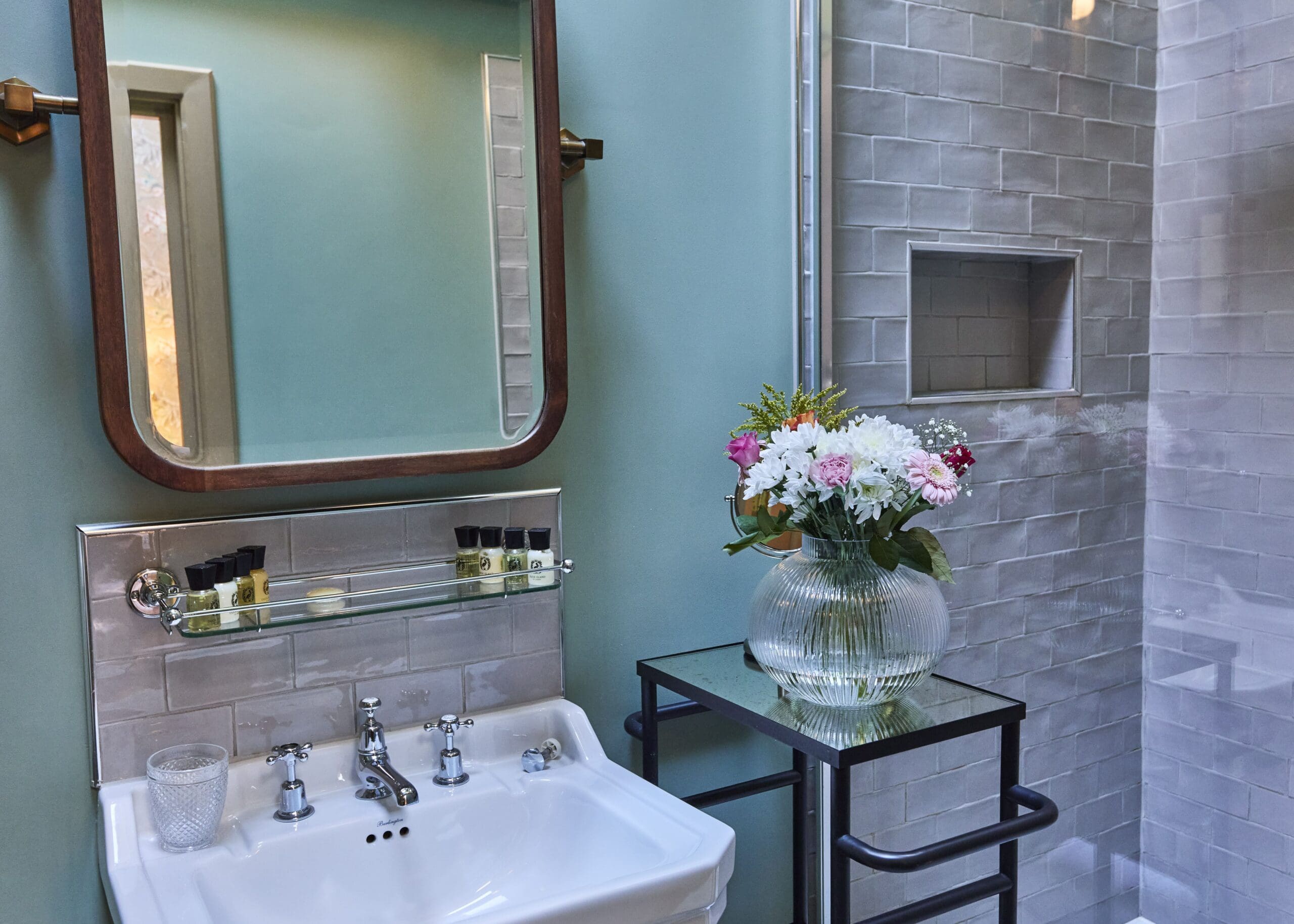 A modern bathroom with a white sink, a large mirror, a glass shelf holding toiletries, and a vase of pink and white flowers on a black table next to a glass shower with light gray tiles.