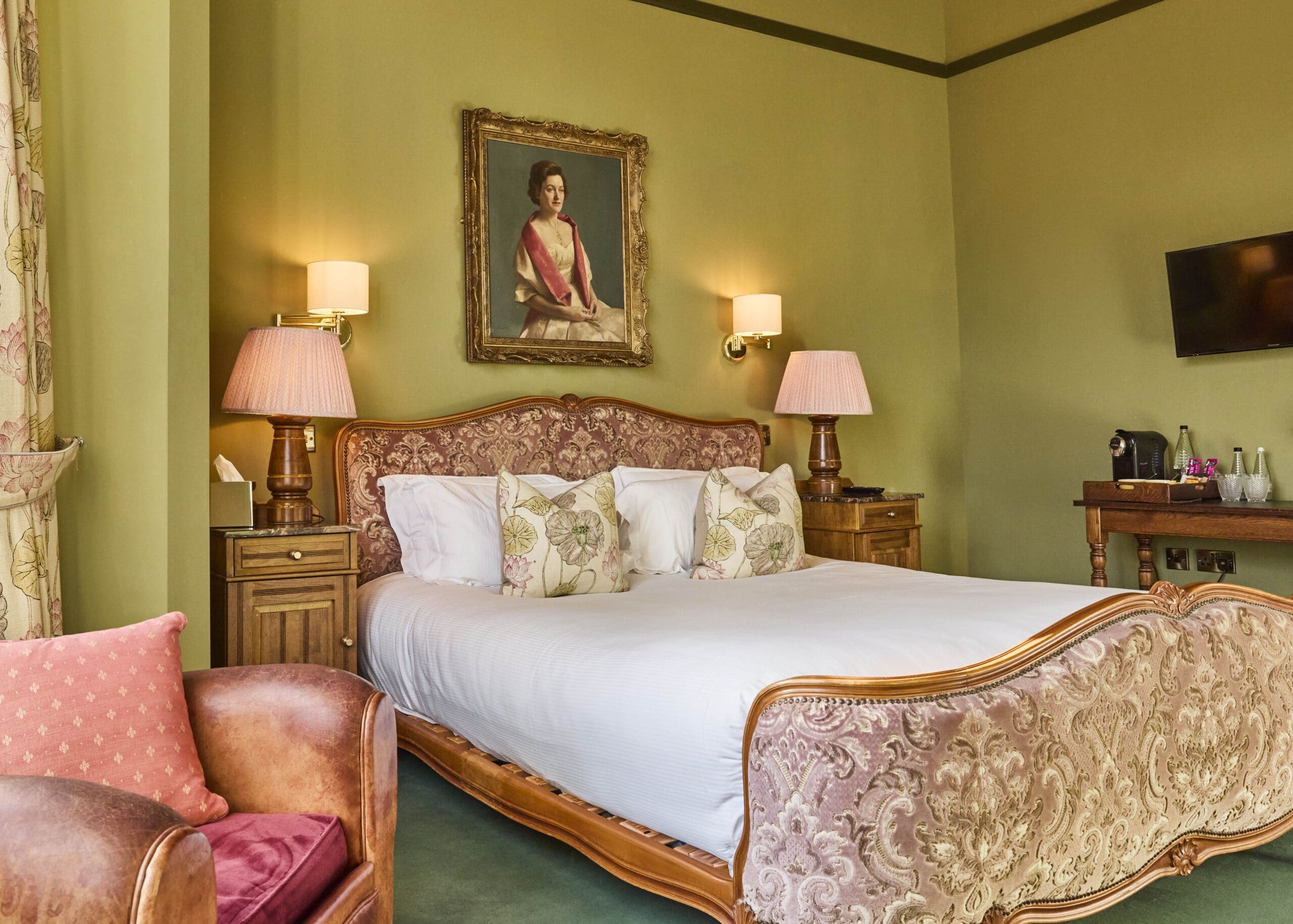 A vintage-style hotel room with a large ornate bed, two bedside tables with pink lamps, a portrait above the bed, an armchair, a flat-screen TV, and a table with a tea set against green walls and carpet.