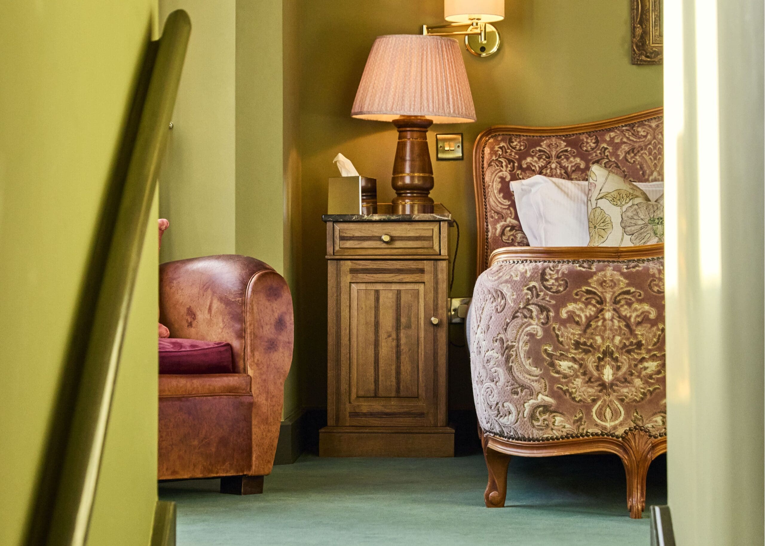 A cozy bedroom features an ornate upholstered bed, a wooden nightstand with a lamp and tissues, and a brown leather armchair, all set against green walls and a green carpet.