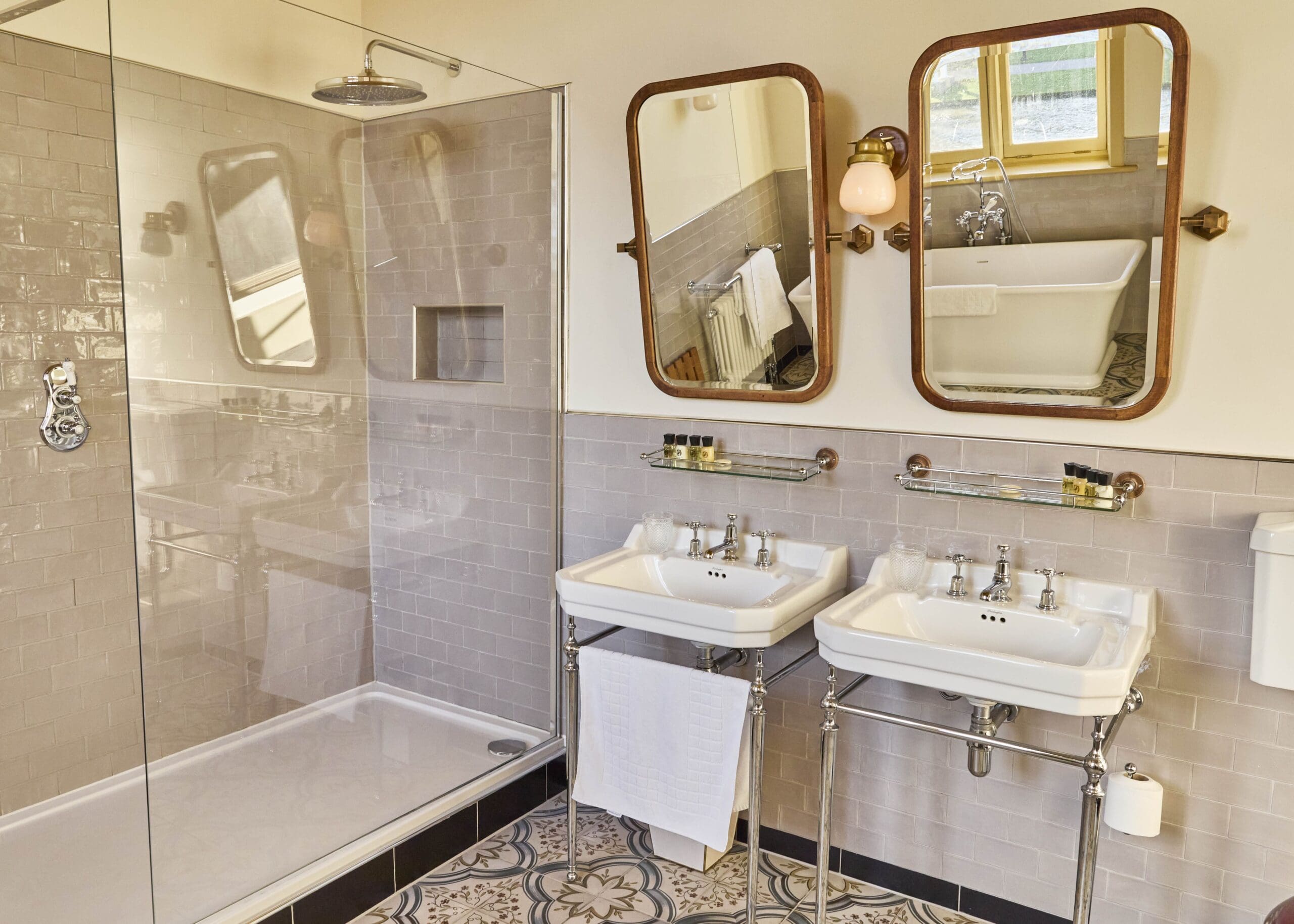 A modern bathroom with a glass shower, two white sinks with mirrors above them, a white towel hanging, decorative floor tiles, and a bathtub near a window in the background.