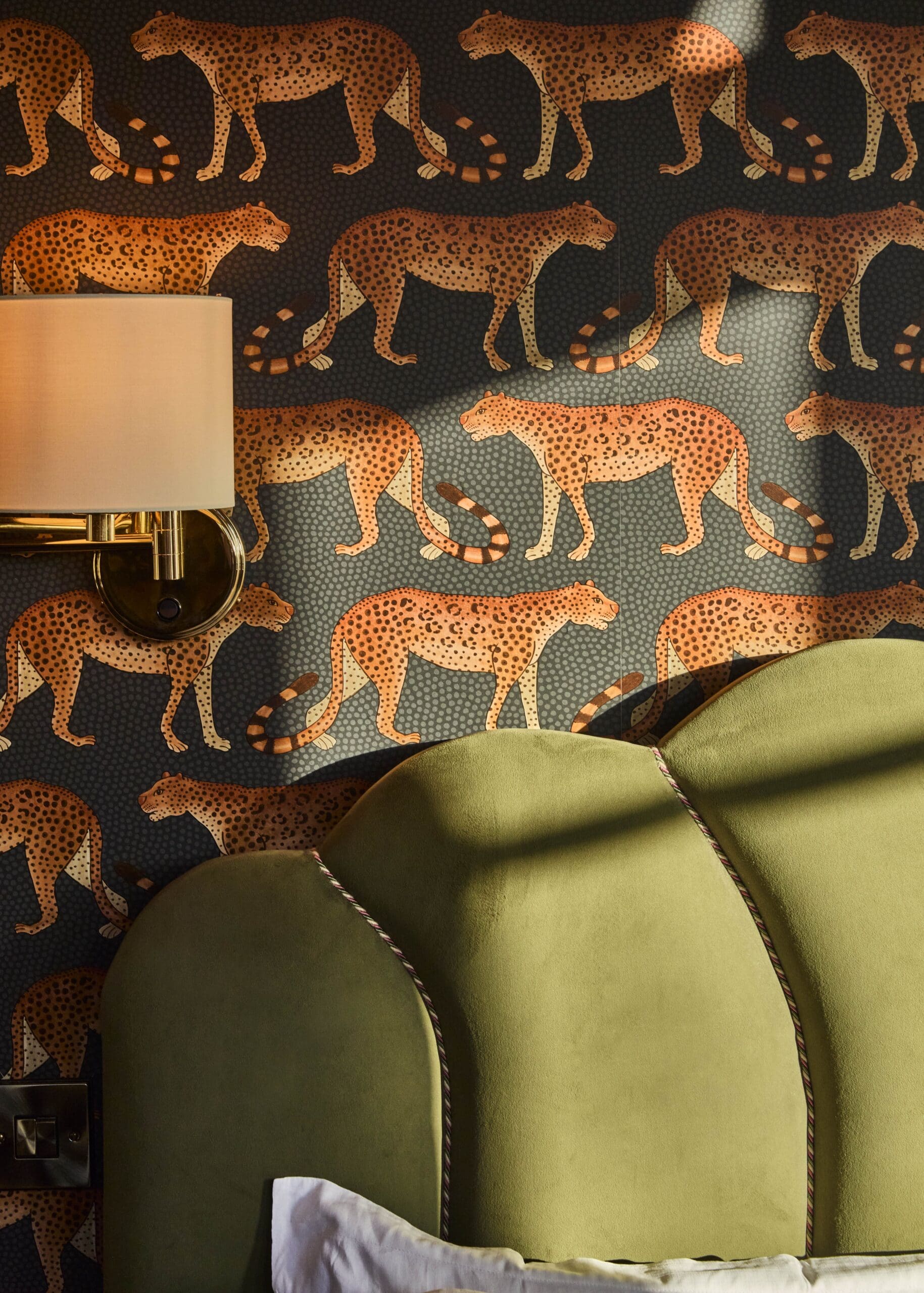 A patterned wallpaper with orange cheetahs on a dark background, partially lit by sunlight, next to a brass wall sconce with a cream shade and an olive green upholstered headboard.