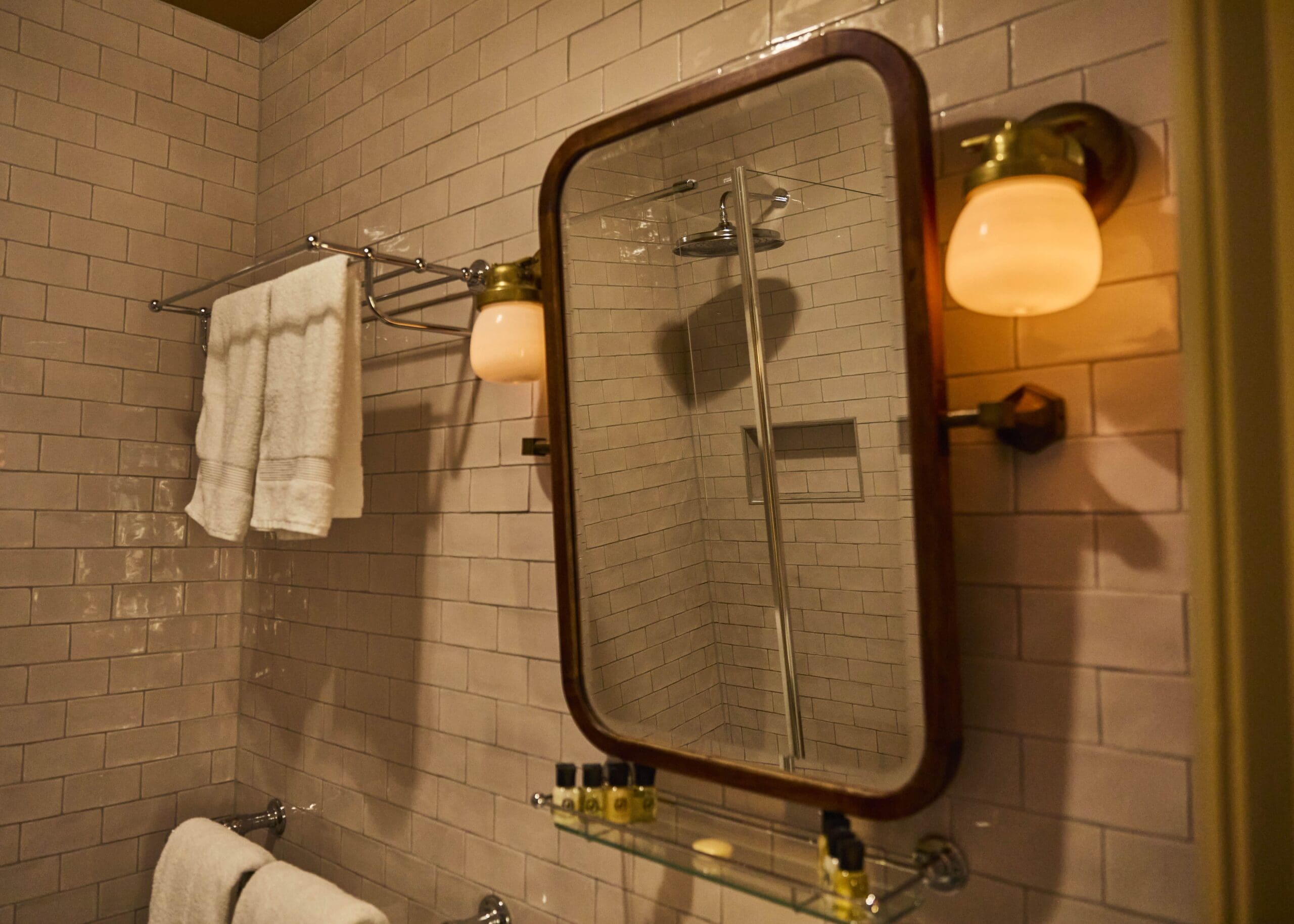 A bathroom with beige subway tile walls, a framed mirror, wall-mounted light fixtures, a towel rack with folded white towels, and a glass shelf holding small toiletries. A shower with a glass door is reflected in the mirror.