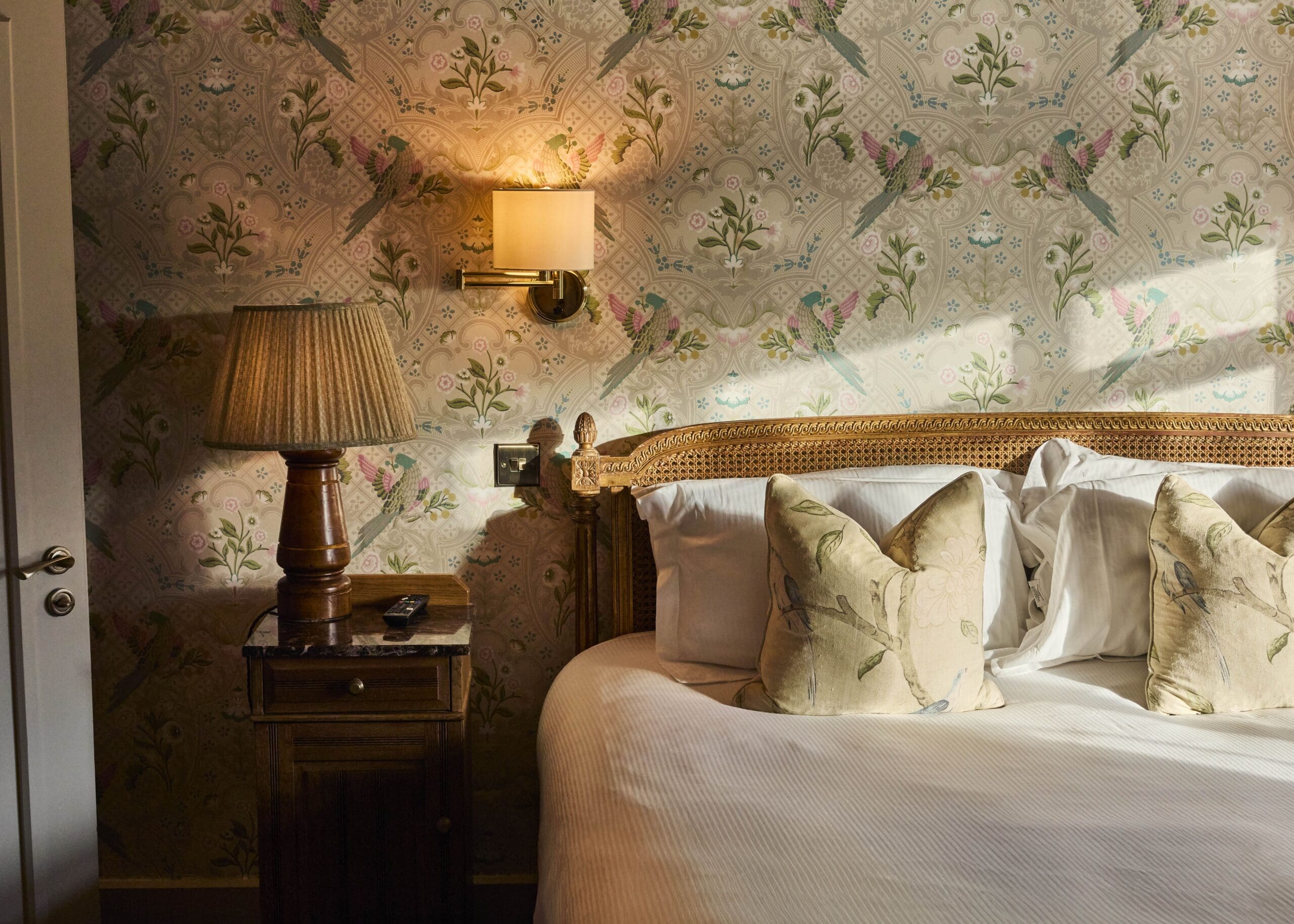 A cozy bedroom with a wooden bed, white bedding, and floral-patterned pillows. A bedside table holds a lamp and remote, and the wallpaper features birds, branches, and flowers. Warm sunlight lights the room.