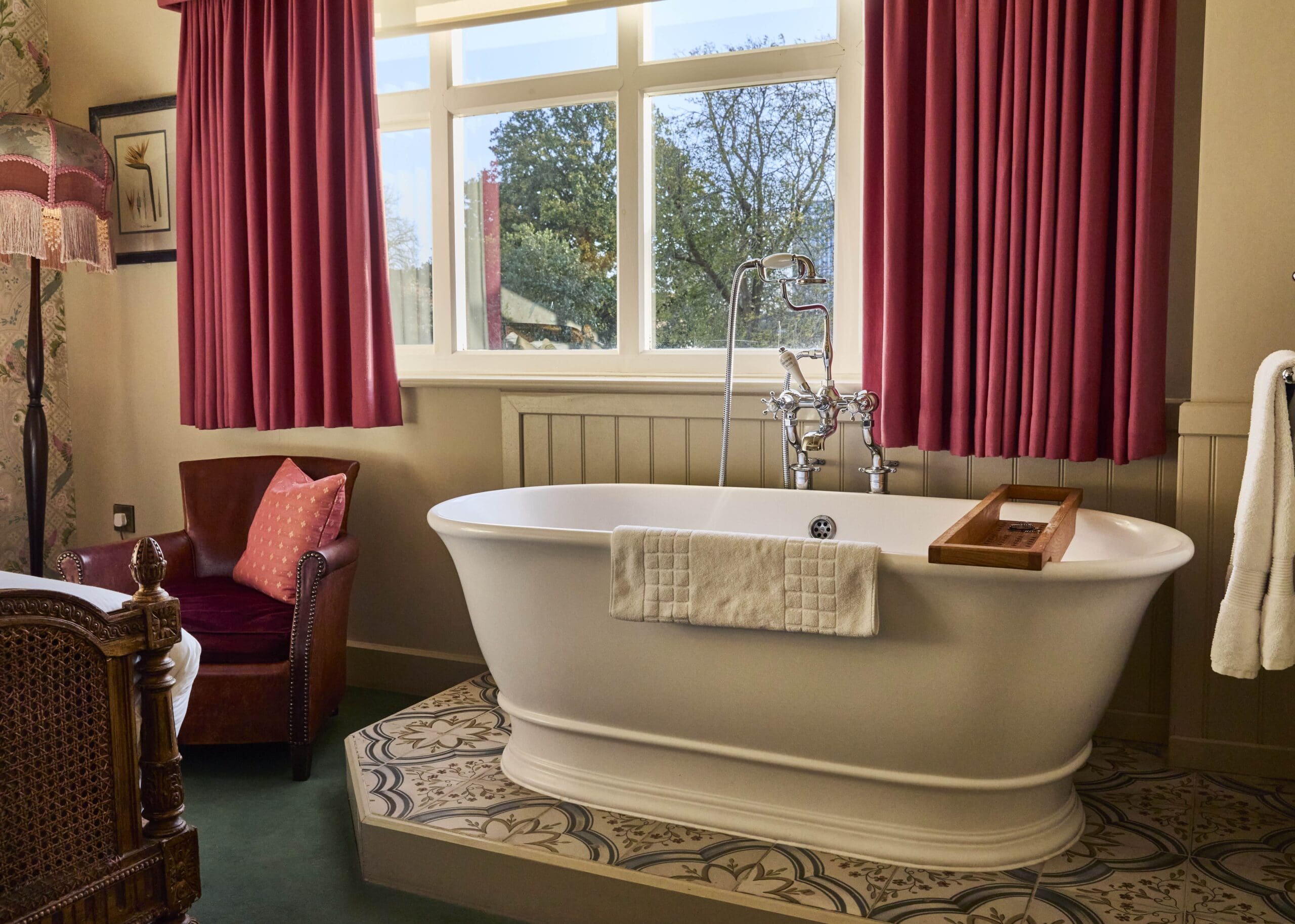 A white freestanding bathtub with a towel draped over the side sits near a window with red curtains. A wooden bath tray rests on the tub. An armchair and lamp are in the corner, and natural light fills the cozy room.