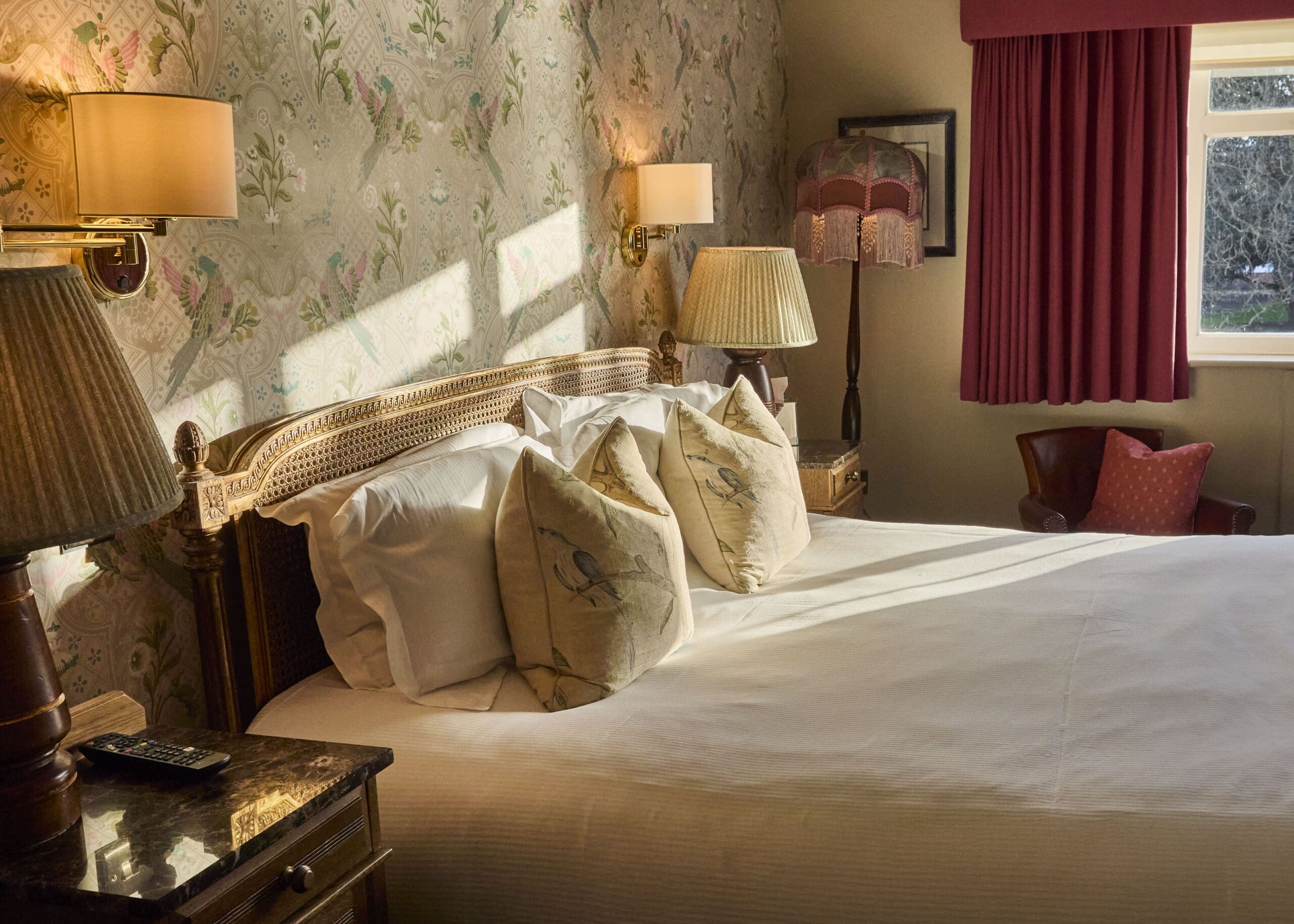 A cozy, sunlit bedroom with a large bed, decorative pillows, classic wooden furniture, floral wallpaper, and a window with red curtains. Lamps and a vintage-style lampshade add a warm, inviting atmosphere.